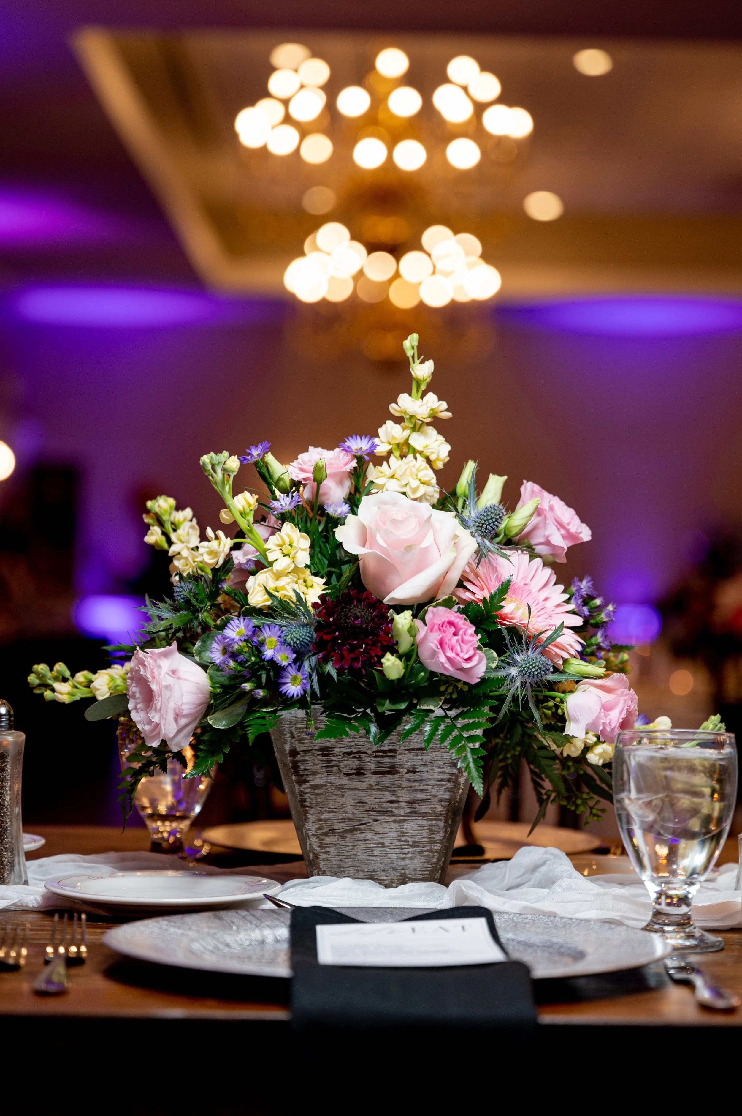 A floral centerpiece with pink roses, purple flowers, and greenery on a table set with plates, silverware, water glasses, and a folded black napkin, in a dimly lit venue with purple and yellow lighting and chandeliers in the background.