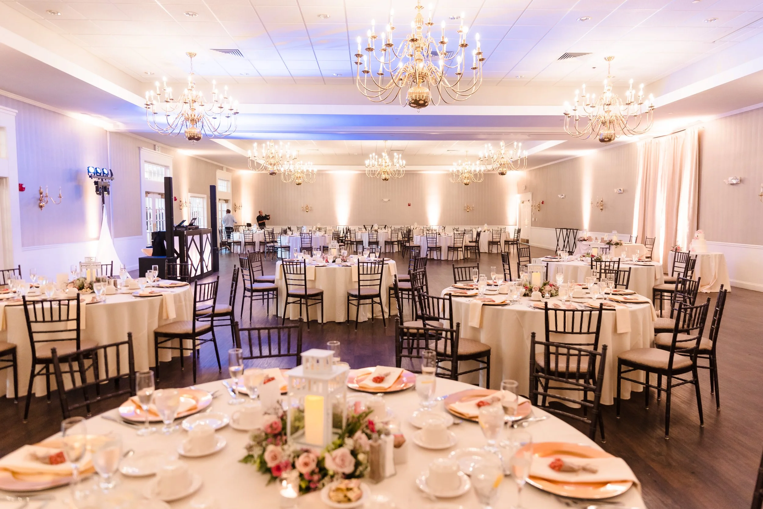 Elegant banquet hall with multiple round tables adorned with white tablecloths, floral centerpieces, and place settings, illuminated by large chandeliers on the ceiling.