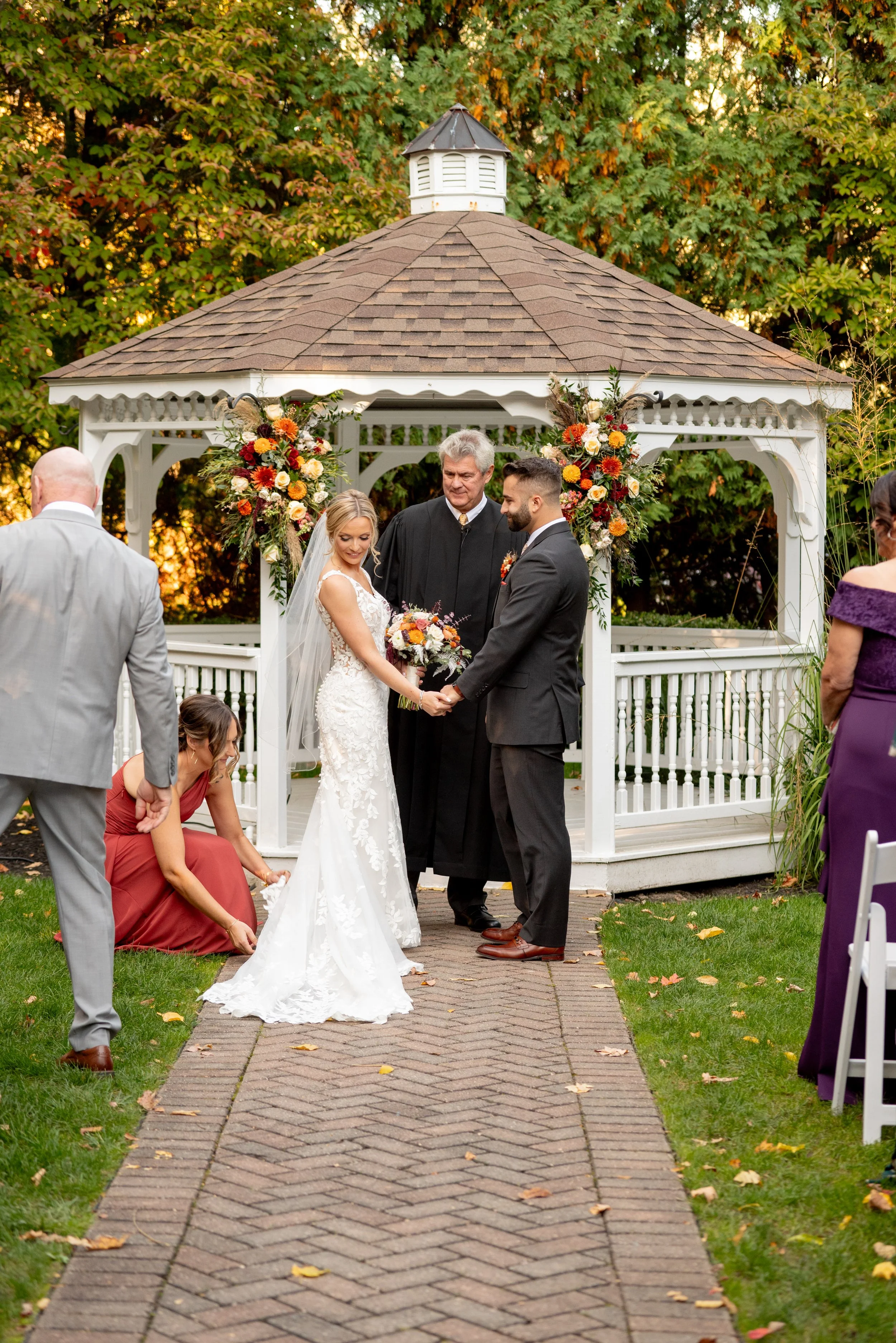 A wedding ceremony outdoors with a bride and groom holding hands, standing under a white gazebo decorated with colorful flowers. An officiant stands between them, and a woman in a rust-colored dress is adjusting the bride's dress at her feet. Guests 