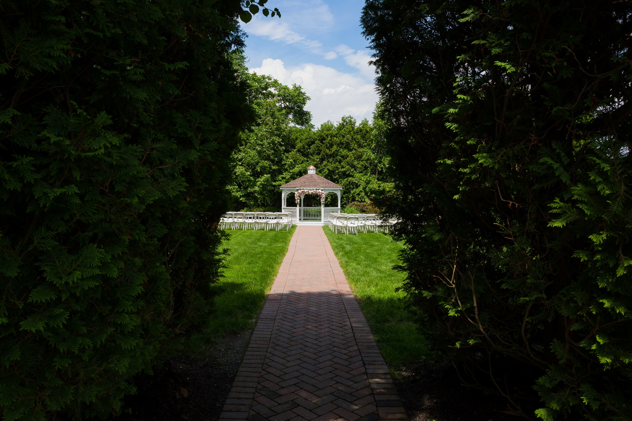 View through a leafy archway leading to a wedding ceremony site with a white gazebo and rows of white chairs on a grassy lawn under a partly cloudy sky.