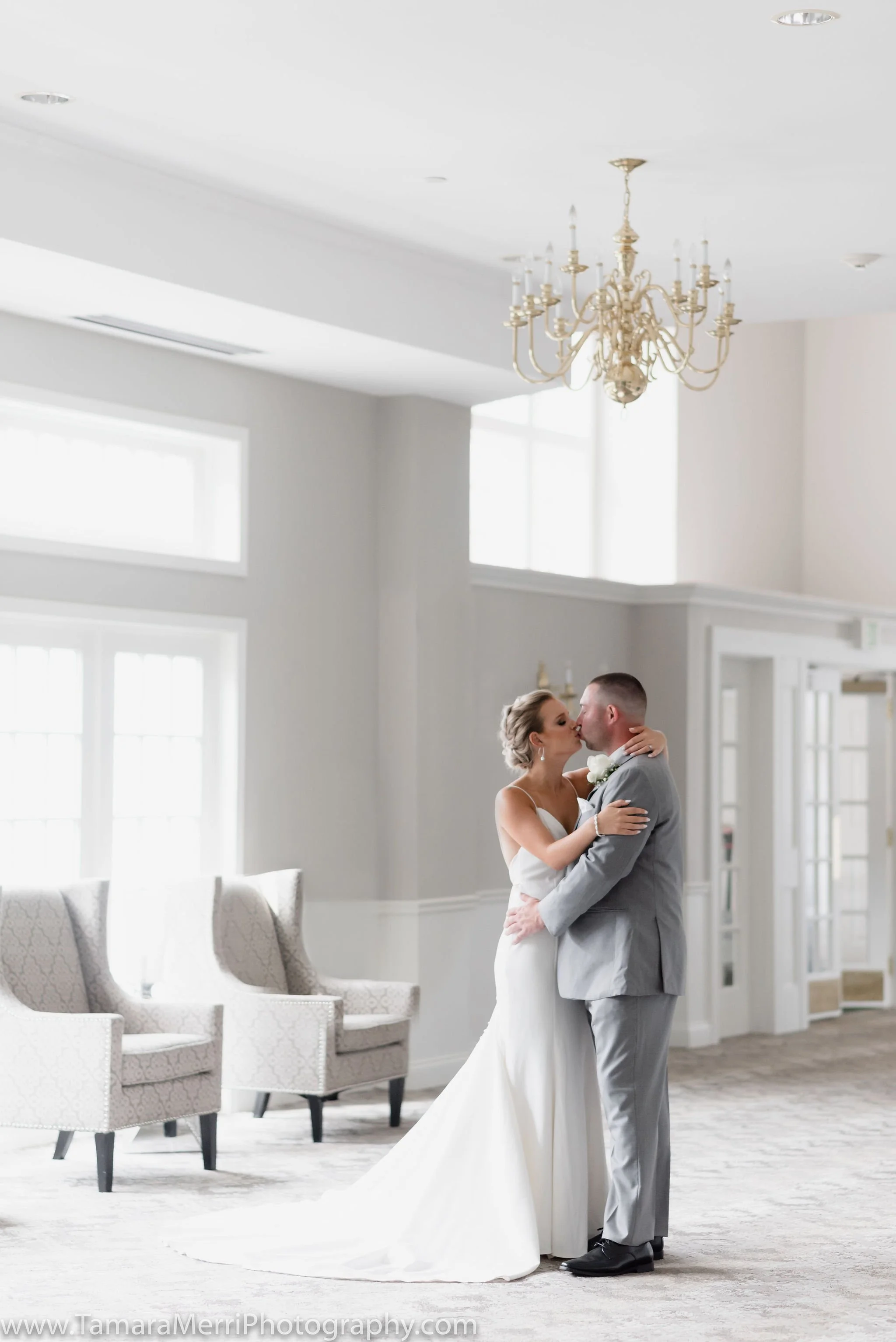 Bride and groom kissing in a bright, elegant room with large windows, white walls, a chandelier, and light-colored chairs.