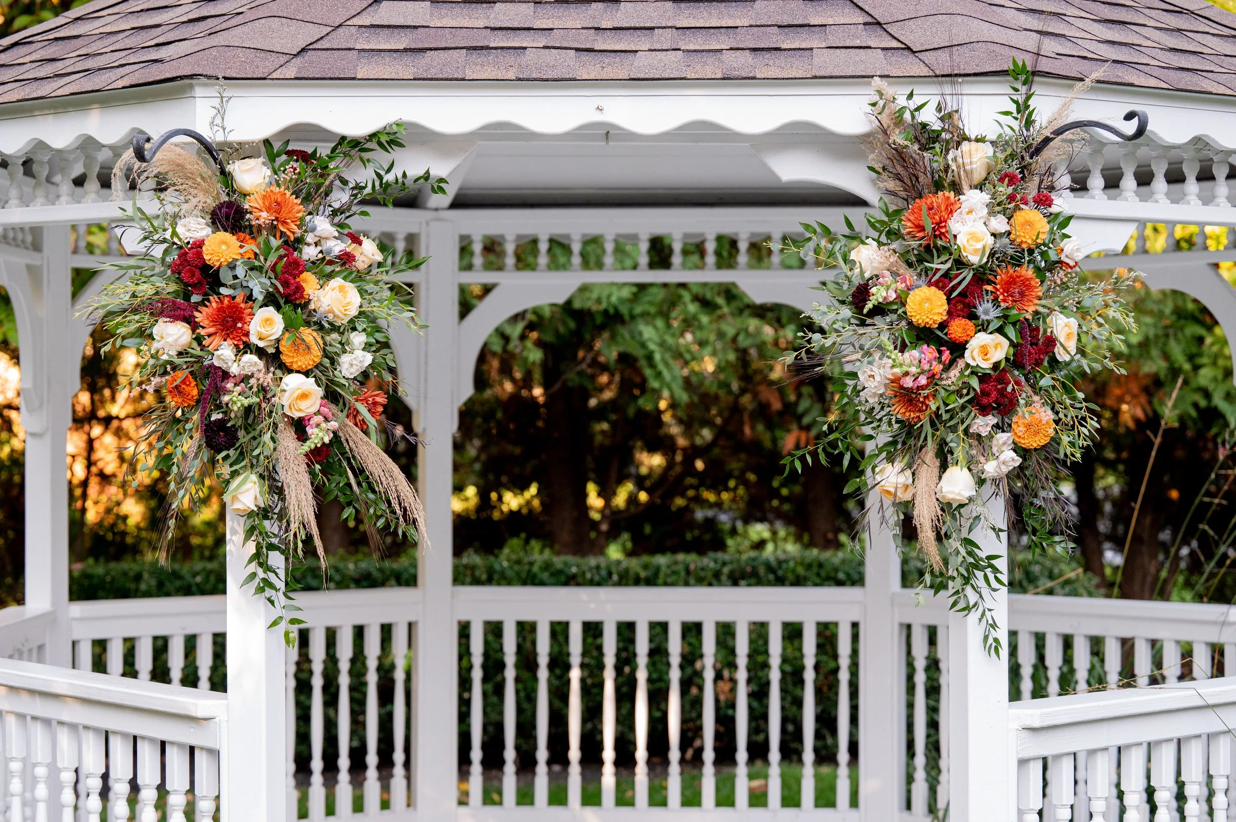Decorative floral arrangements on a white gazebo with a shingled roof, set outdoors with trees in the background.