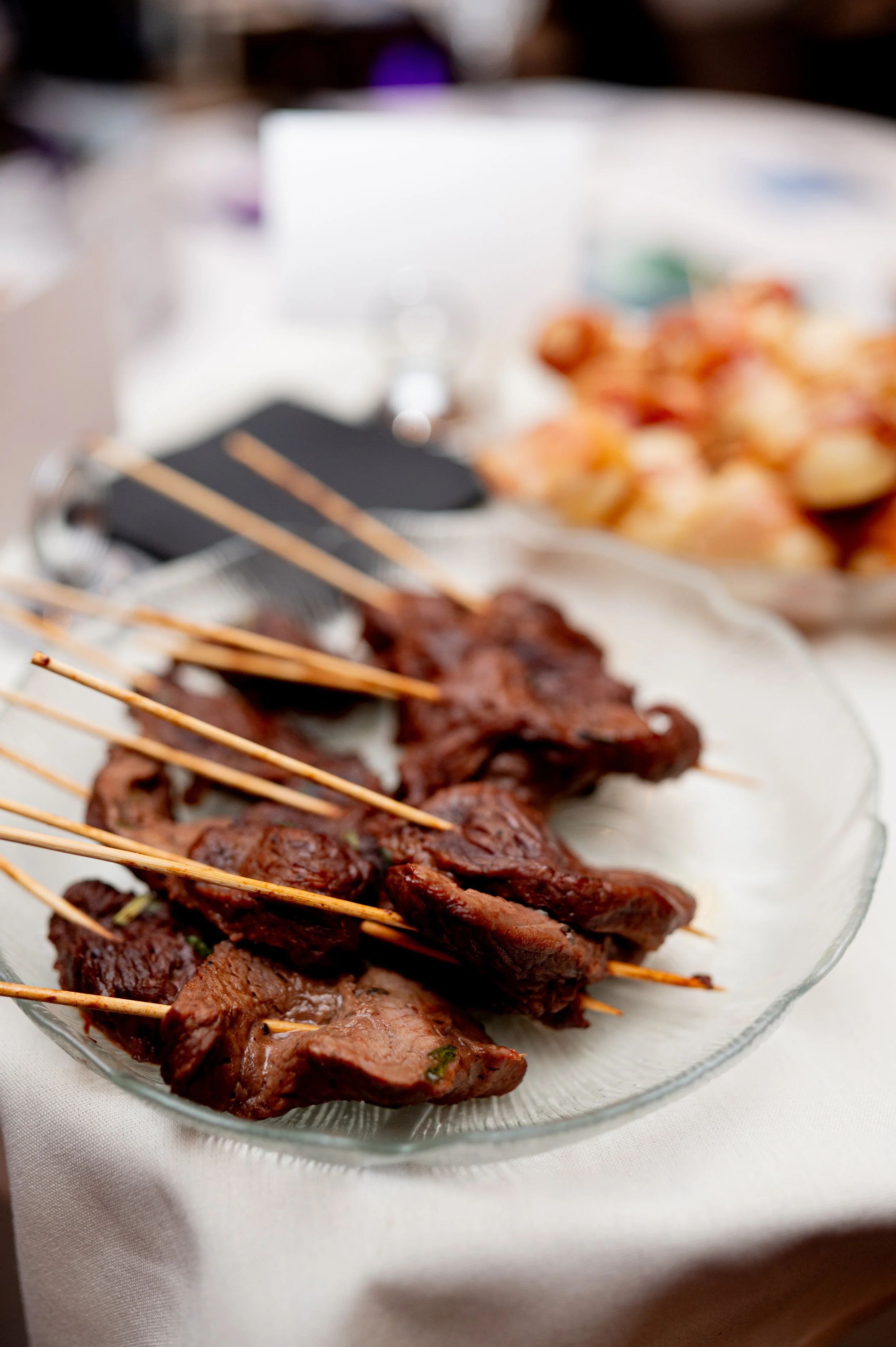 Skewered grilled meat on a glass platter at a table, with blurred background including a bowl of bread rolls.