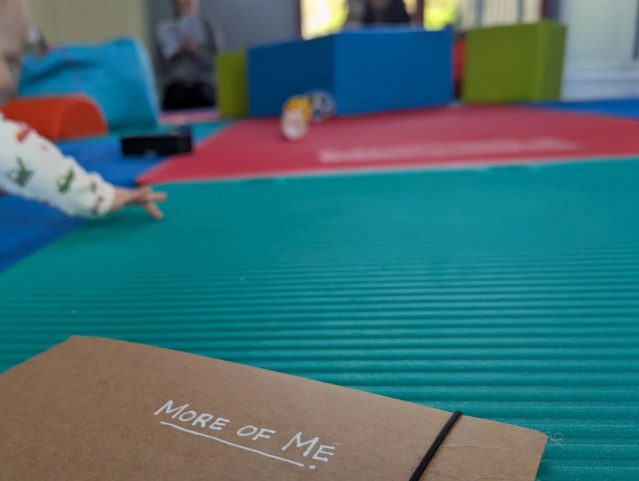 Colorful foam mats on the floor in a play area with children in the background, and a brown notebook with the title "More of Me" in the foreground.