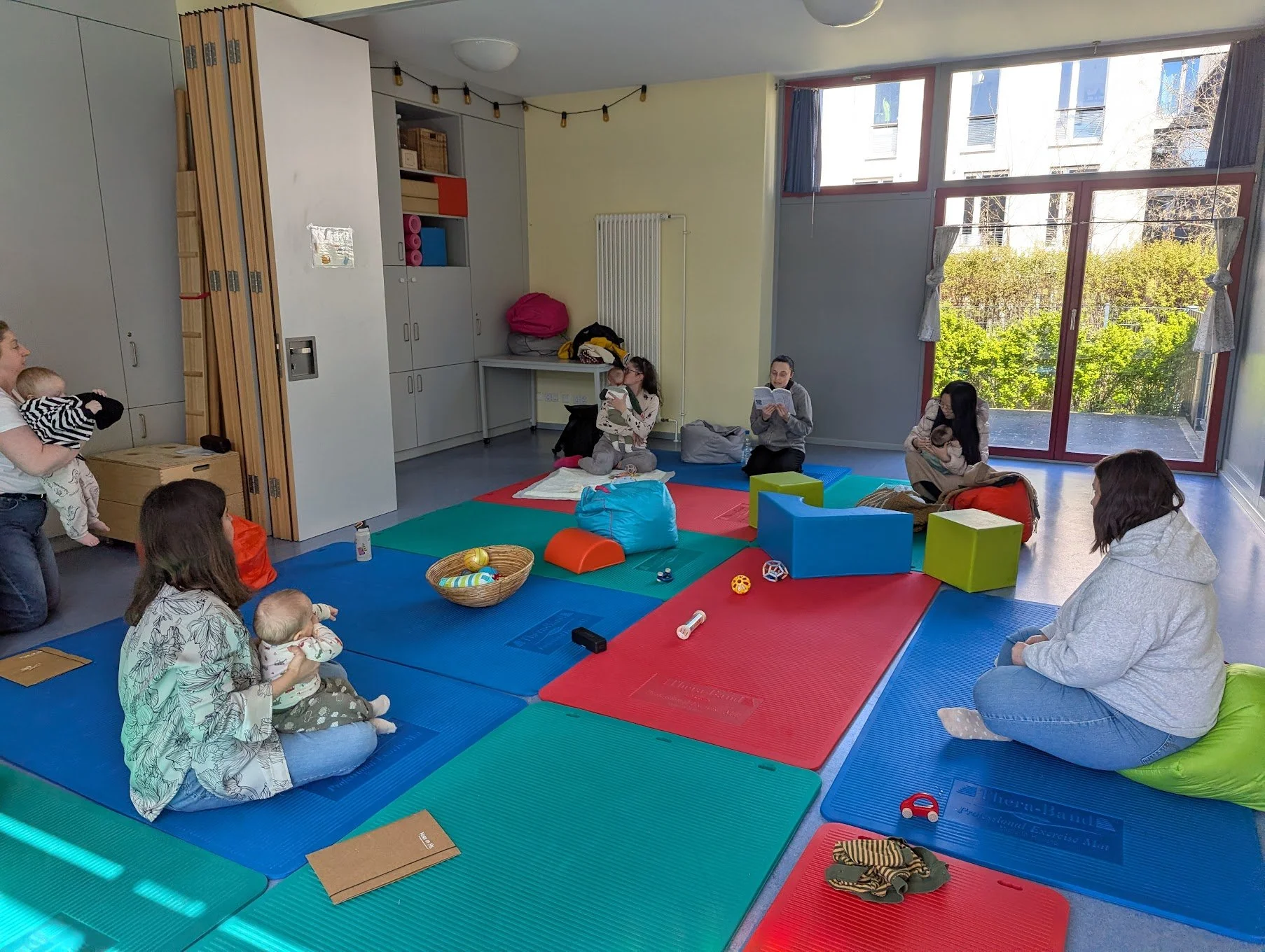 A group of women and children sitting on colorful mats in a bright room with large windows and green trees outside. Some women are holding babies, and some are reading or sitting quietly. Toys and bags are scattered around.