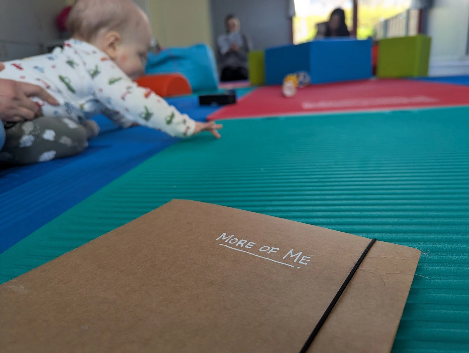 A brown notebook labeled "More of Me" resting on a multicolored padded surface at a play area, with a baby reaching out in the background.