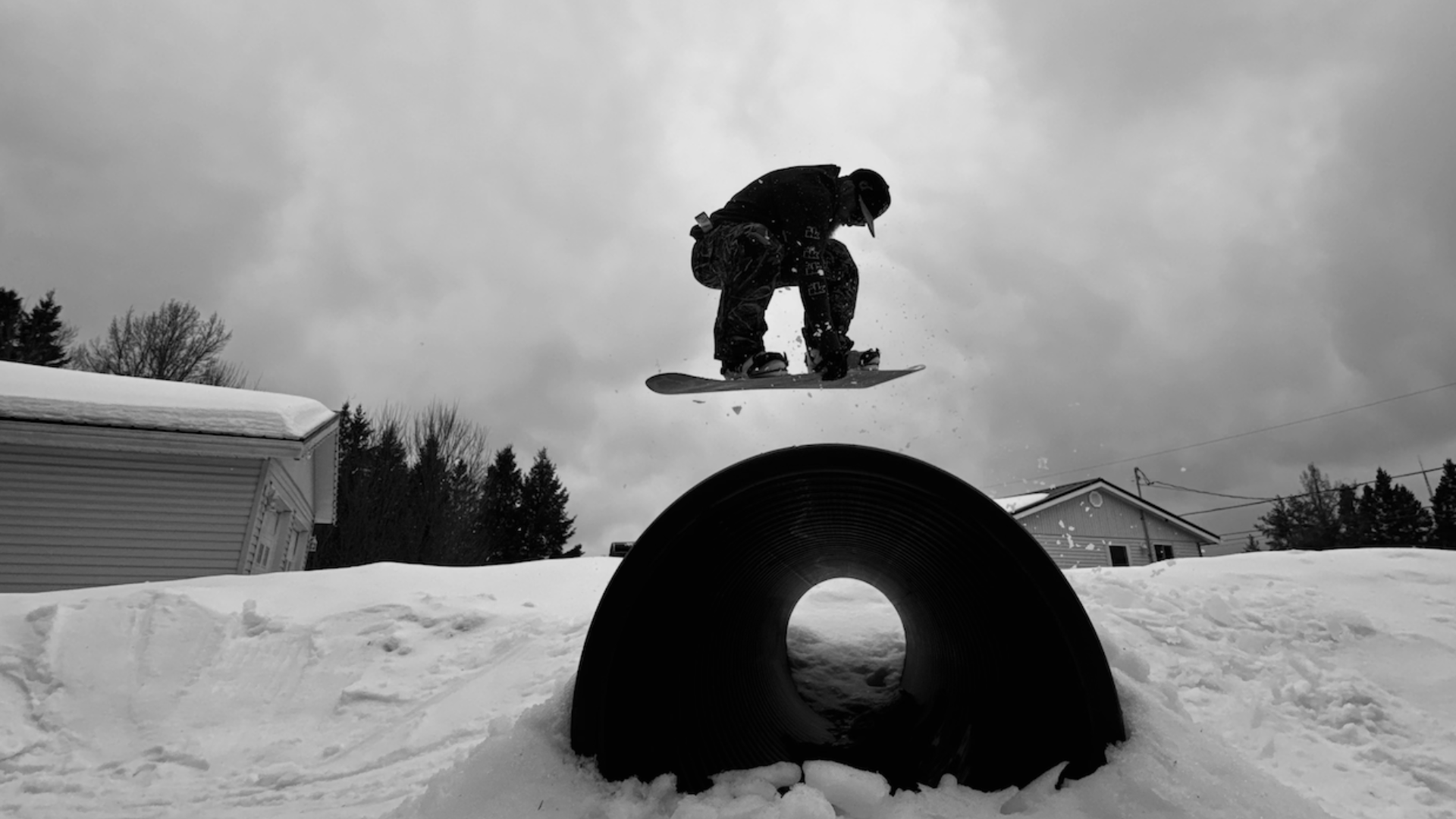 A person in winter clothing snowboarding in mid-air over a large pipe in a snowy landscape with houses and trees in the background, under cloudy sky.