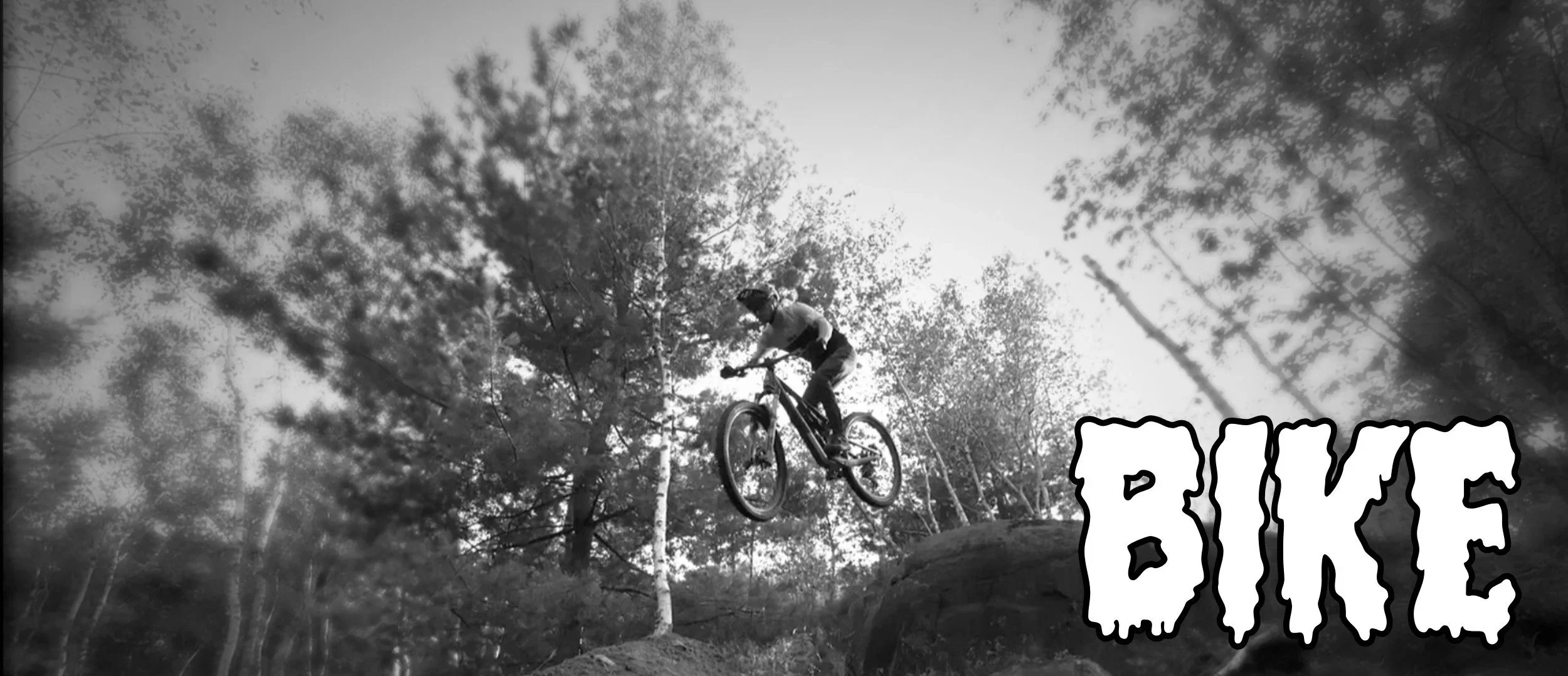Black and white photo of a mountain biker mid-air jumping off a dirt ramp in a forested area. The sky is visible through the trees. The word 'BICE' appears in large, stylized font in the lower right corner.