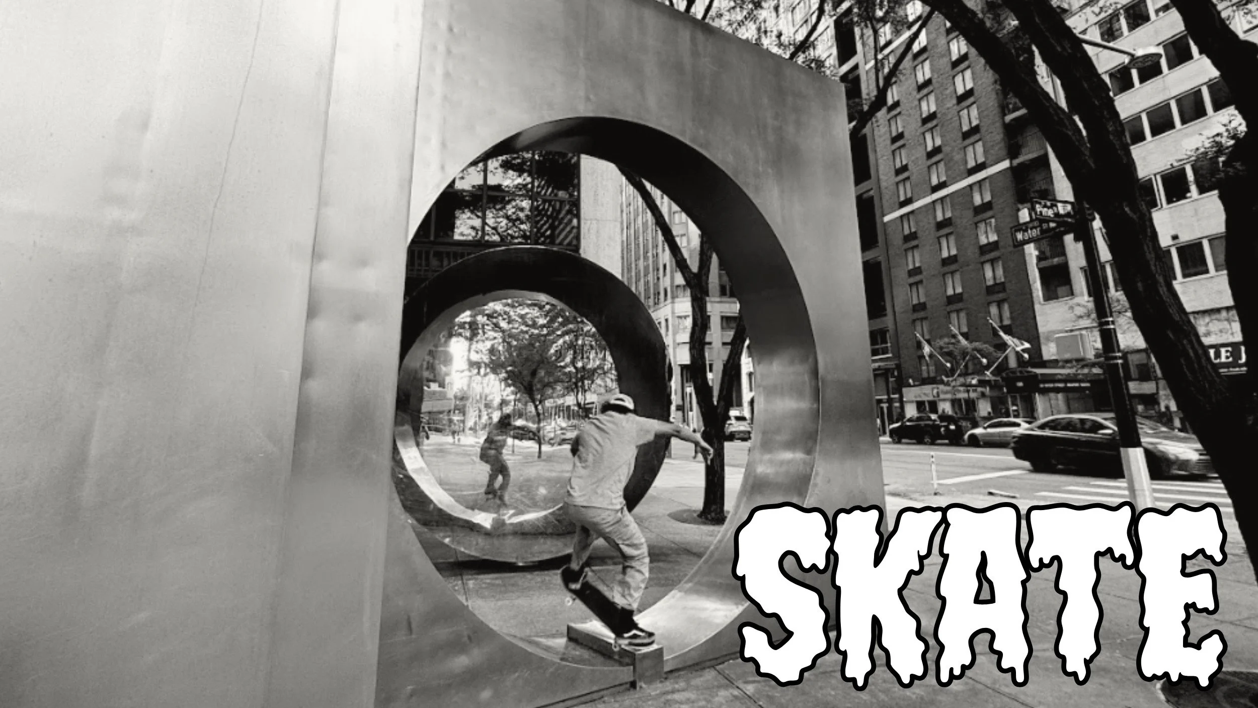 Black and white photo of two people outside near a large, circular metallic sculpture in an urban setting, with tall buildings and trees in the background.