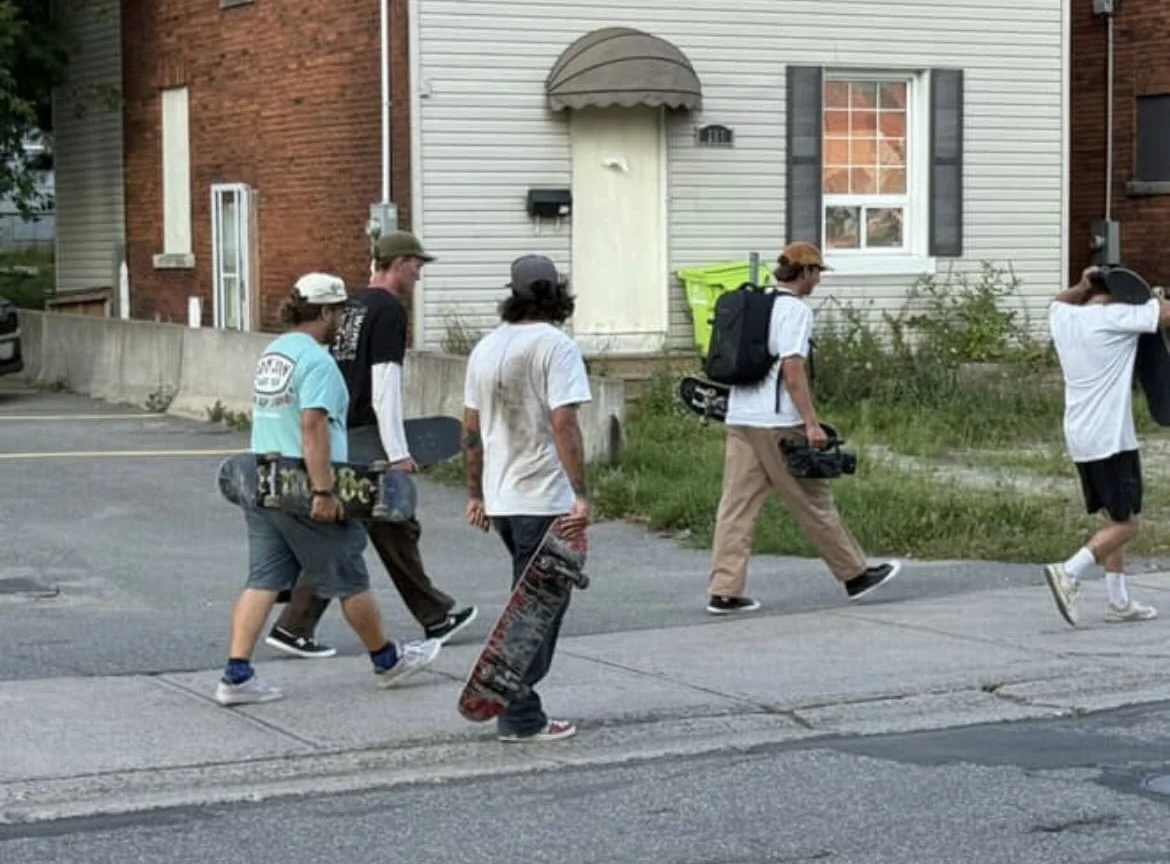Five skaters walking on the street, each carrying skateboards and some with backpacks.