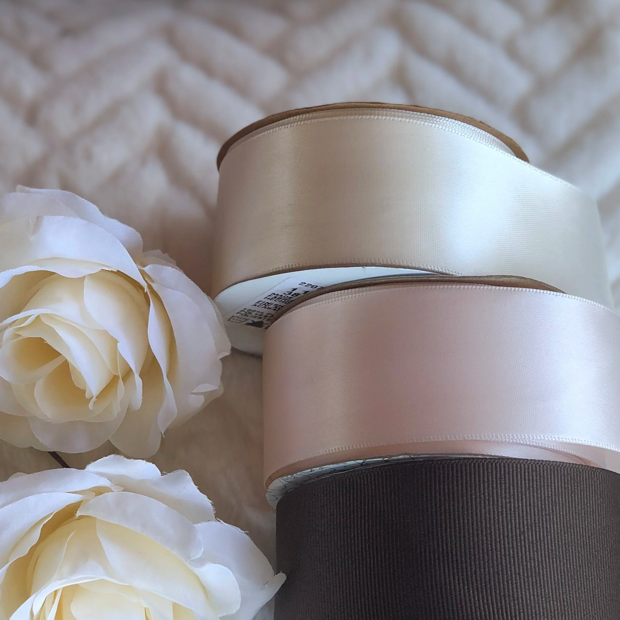 Three rolls of satin ribbon in cream, blush, and dark brown sit next to two cream-colored roses on a soft, cream fabric background with herringbone pattern.