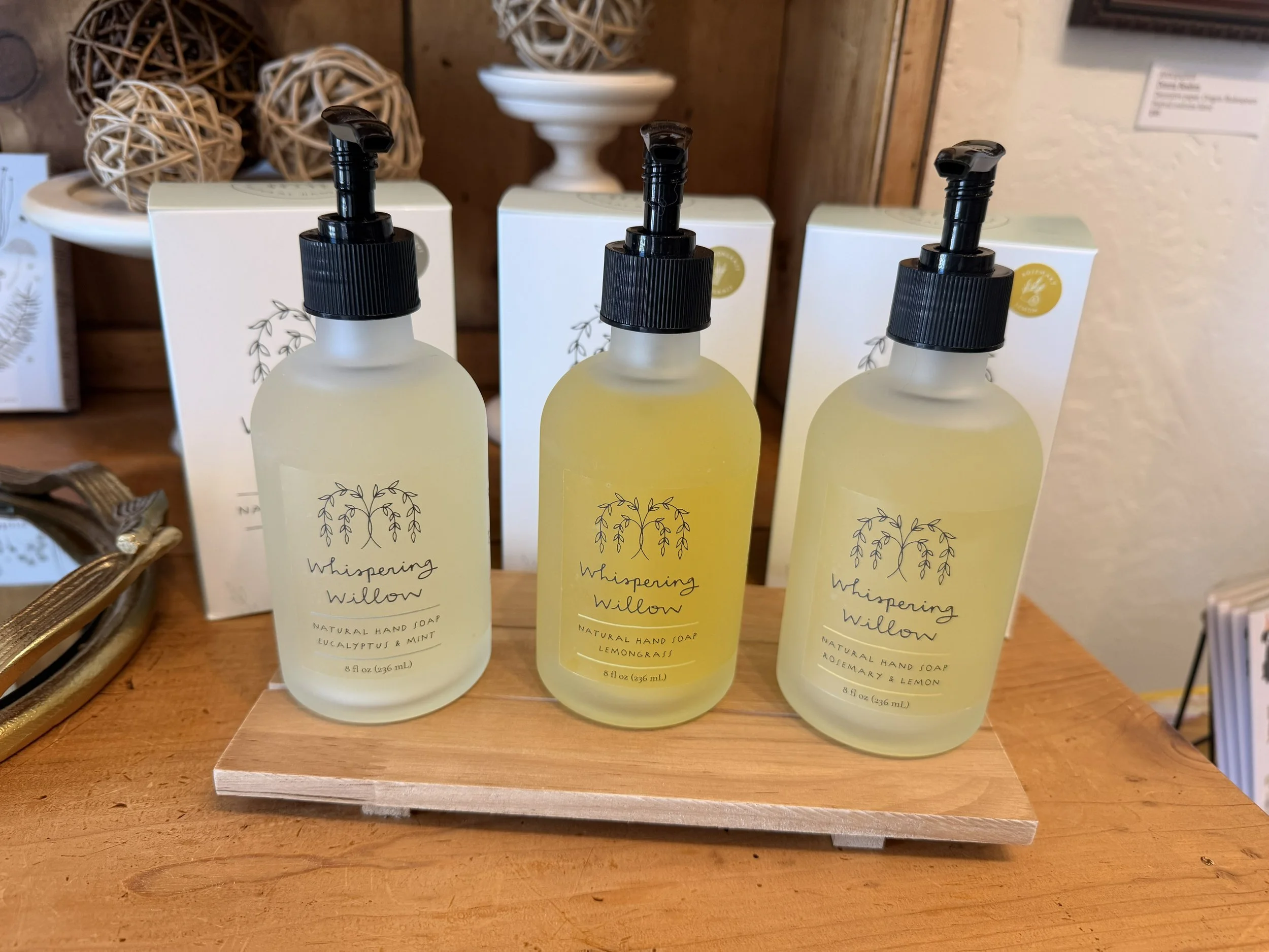 Close-up of Whispering Willow's Lemongrass Natural Pump Hand Soap on display with two other bottles at Bloomie's Boutique