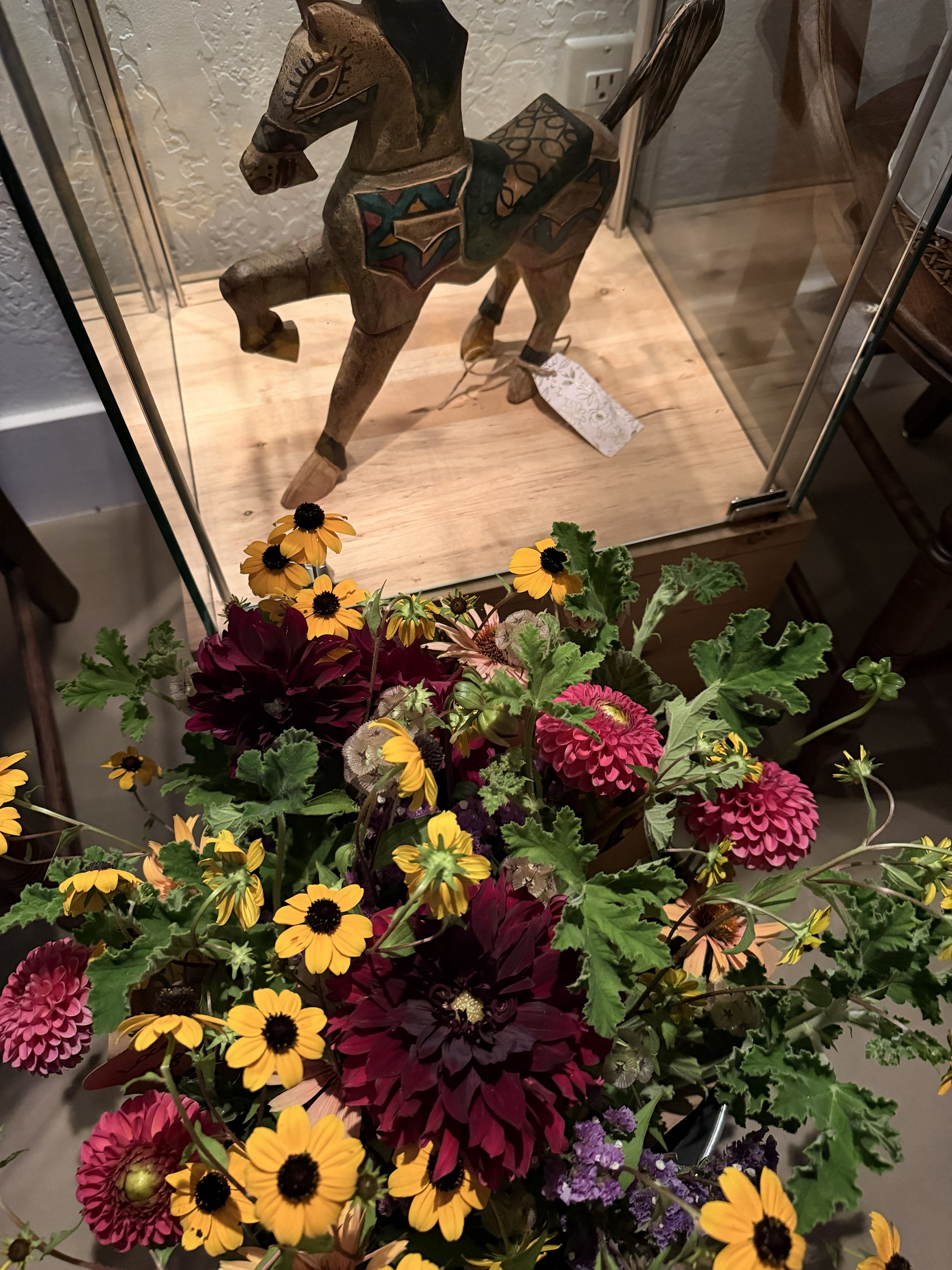 Upcycled folk art horse in display case at Bloomie's Boutique with flowers in evening light