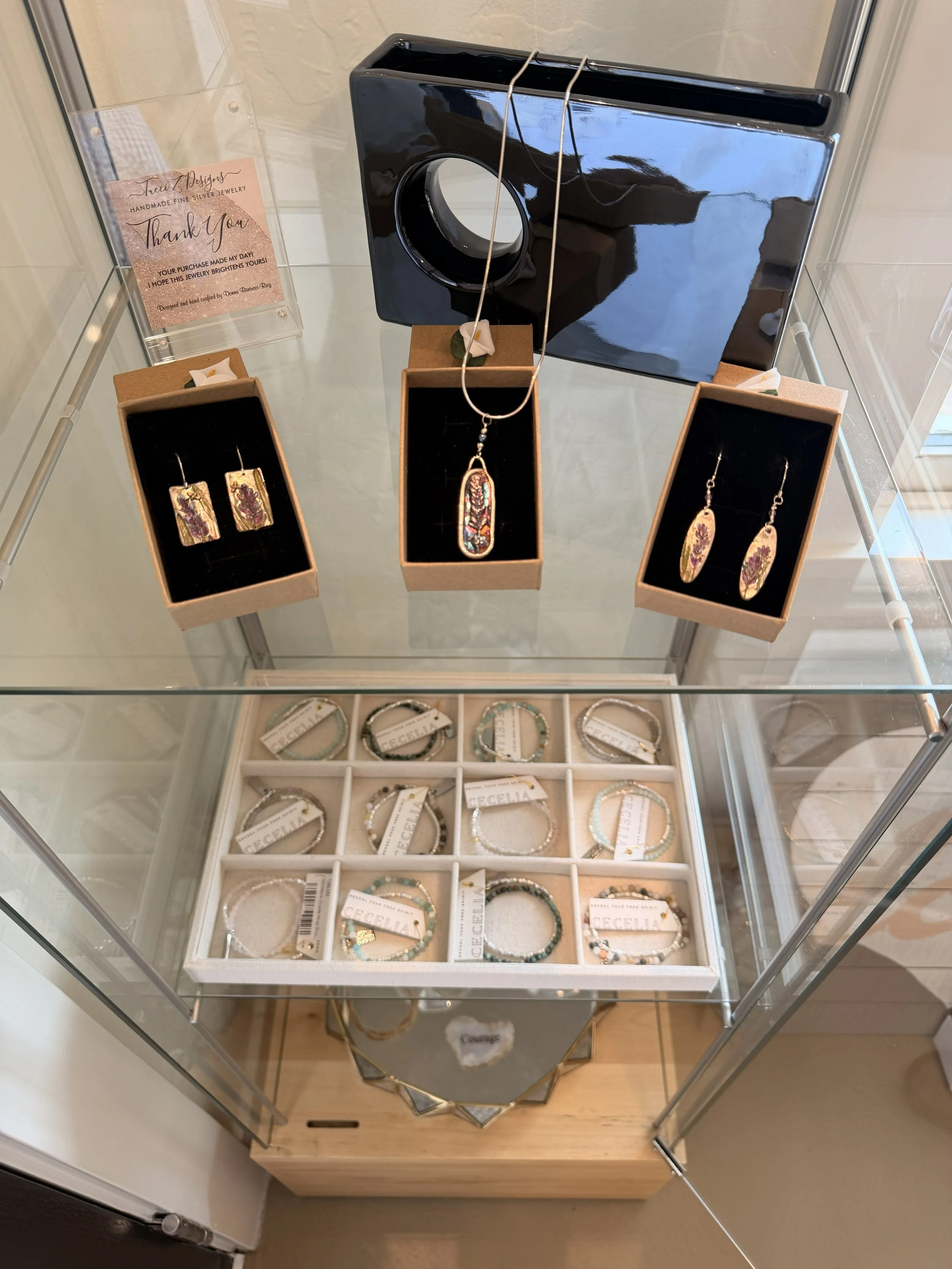 Tucci'Z Designs fine silver pendant with lavender motif on display along with fine silver earrings in display case at Bloomie's Boutique. Bracelets by Cecelia Designs on display below.