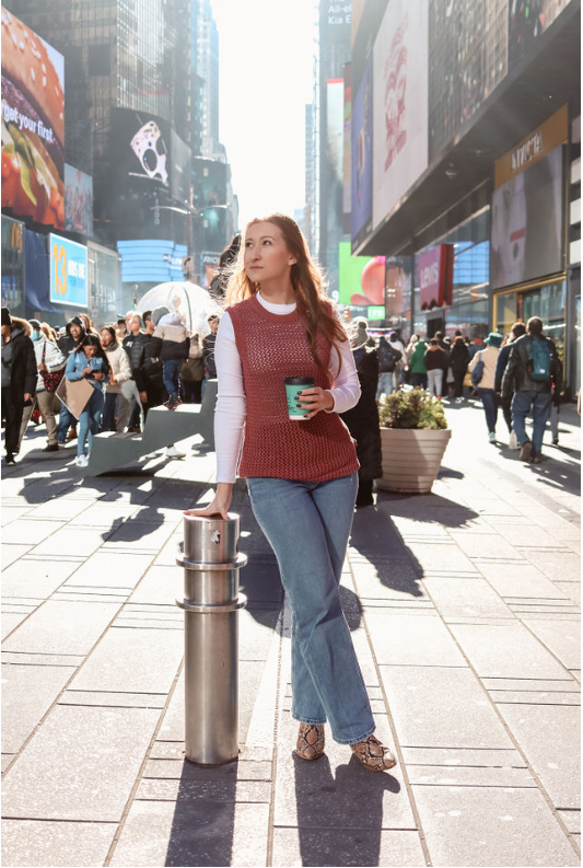 A woman standing on a busy city street holding a coffee cup and leaning on a metal post, with digital billboards and crowds in the background.