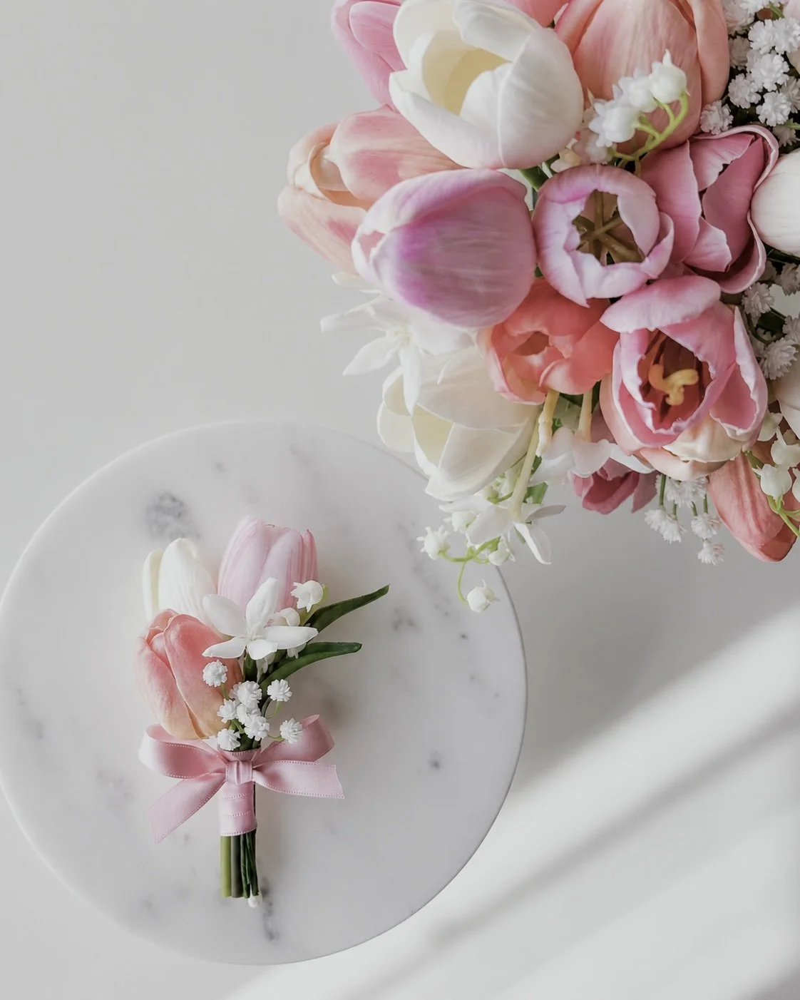 A floral boutonniere with pink tulips, white lily-like flowers, and small white filler flowers, tied with a pink ribbon, placed on a white marble surface. A bouquet of pink and white flowers is seen in the background.