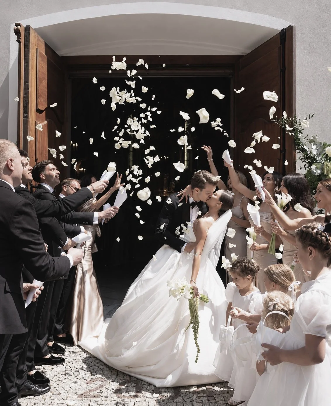 A newlywed couple kisses as they walk out of a church, surrounded by friends and family throwing flower petals. The bride is holding a bouquet of white flowers, and everyone is dressed in formal wear for a wedding celebration.