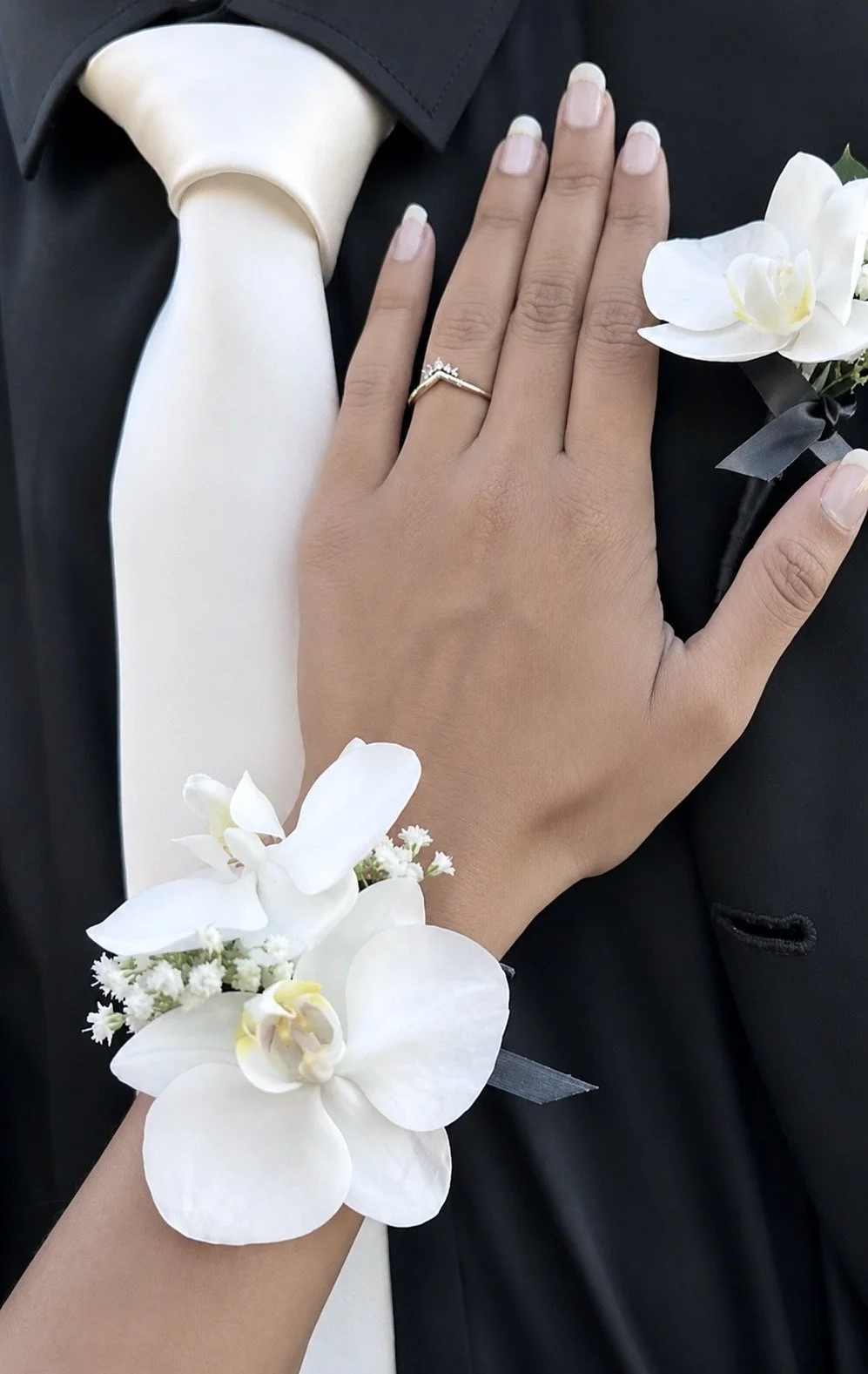 Close-up of a woman's hand with a ring on her finger, resting on a man's chest. The woman is wearing a flower corsage with white orchids and baby's breath on her wrist. The man is in a black suit with a white shirt and a silver tie, and there is a white orchid boutonniere on his lapel.