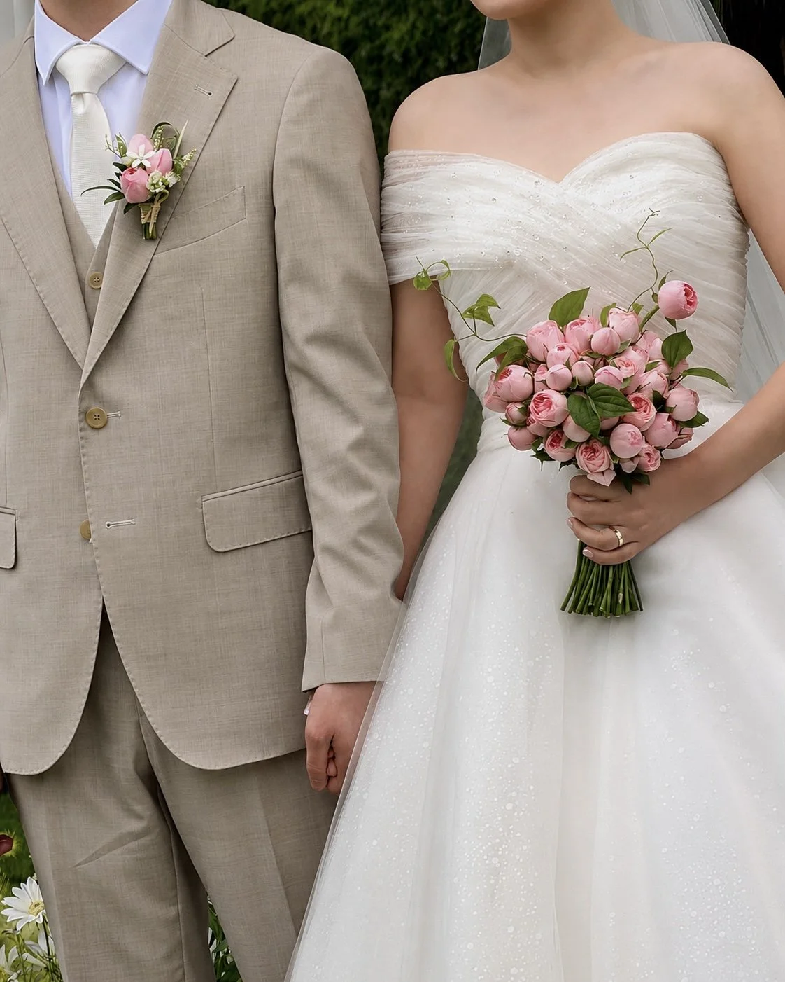 A bride holding a pink rose bouquet and a groom in a beige suit standing side by side at a wedding.