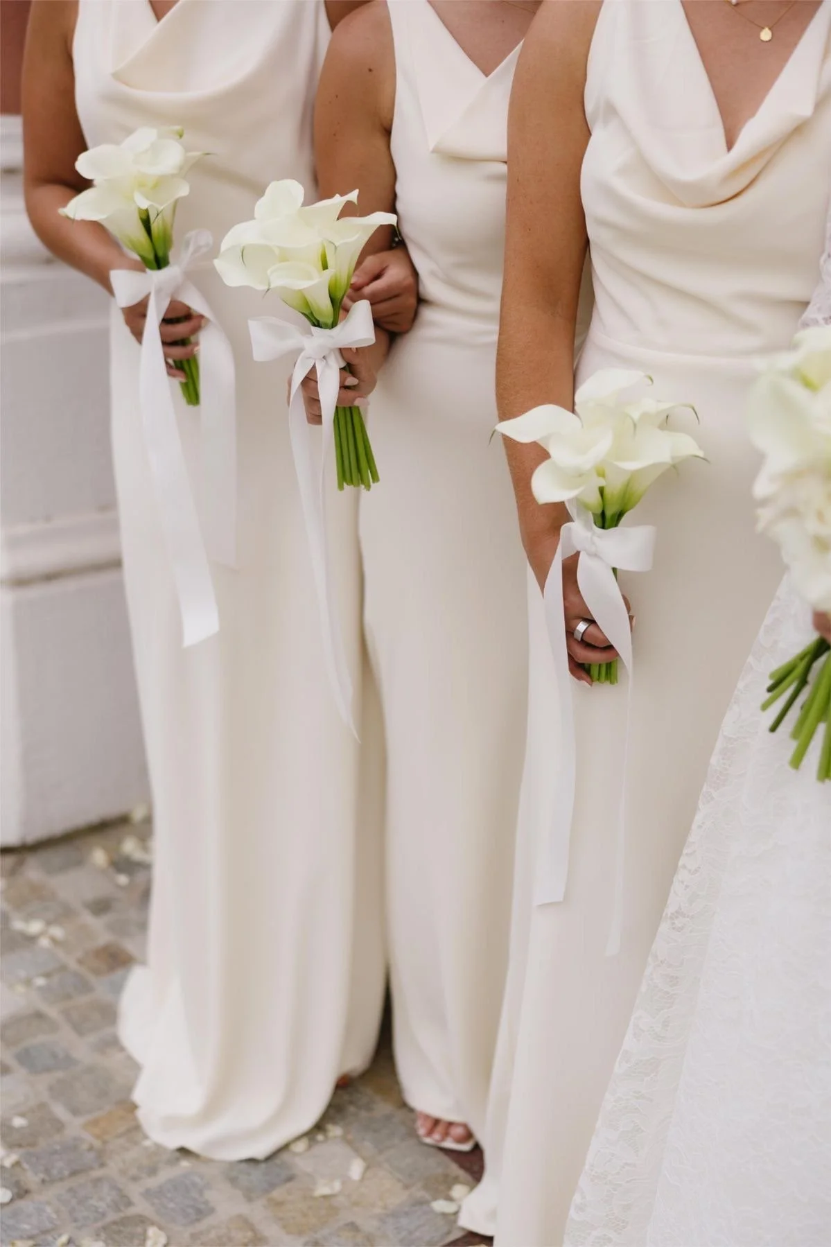 Women dressed in white, holding bouquets of white calla lilies tied with white ribbons, at a wedding ceremony.
