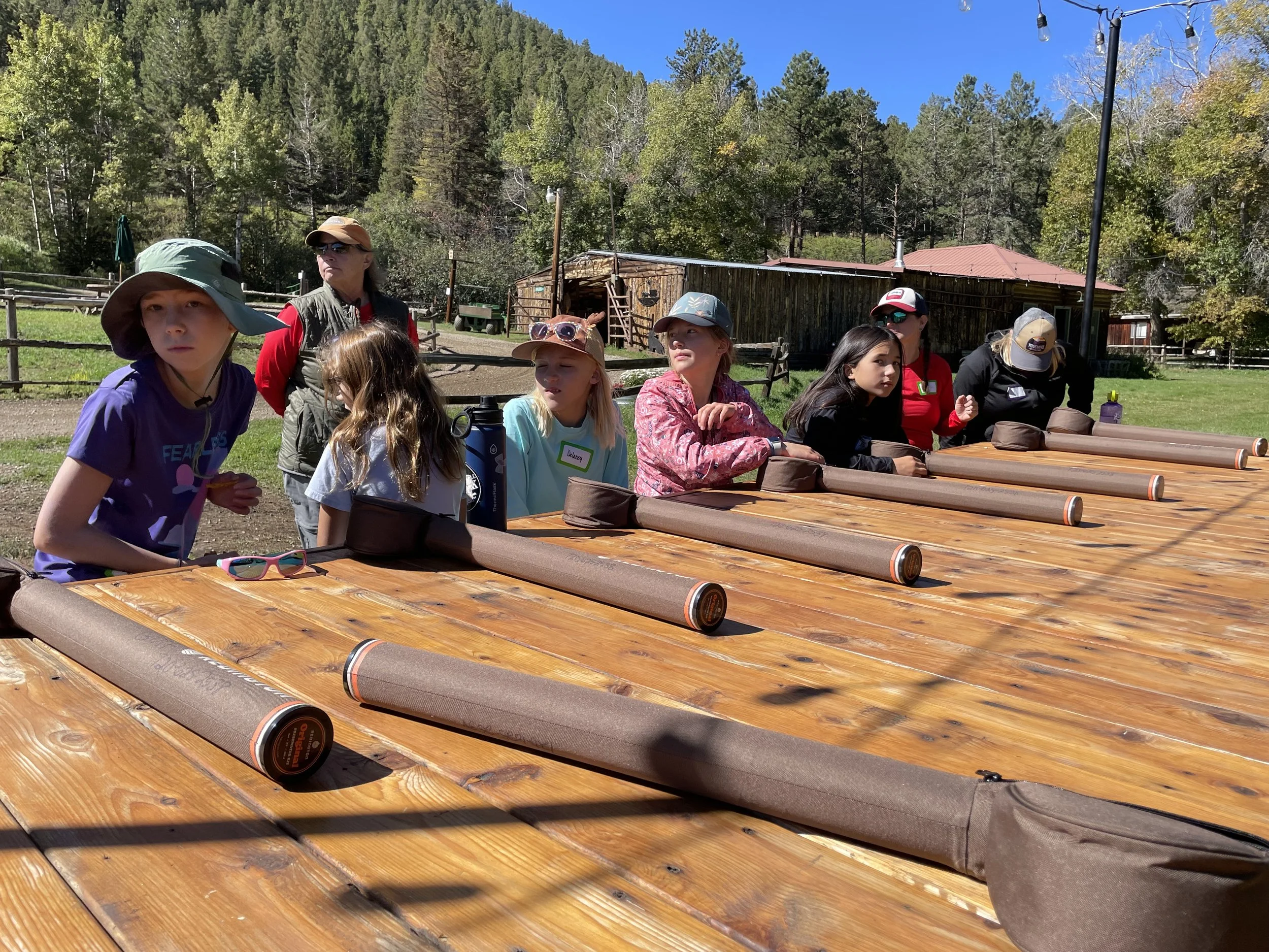 Group of children and adults at an outdoor archery range with wooden targets, surrounded by trees and a rustic building under a clear blue sky.