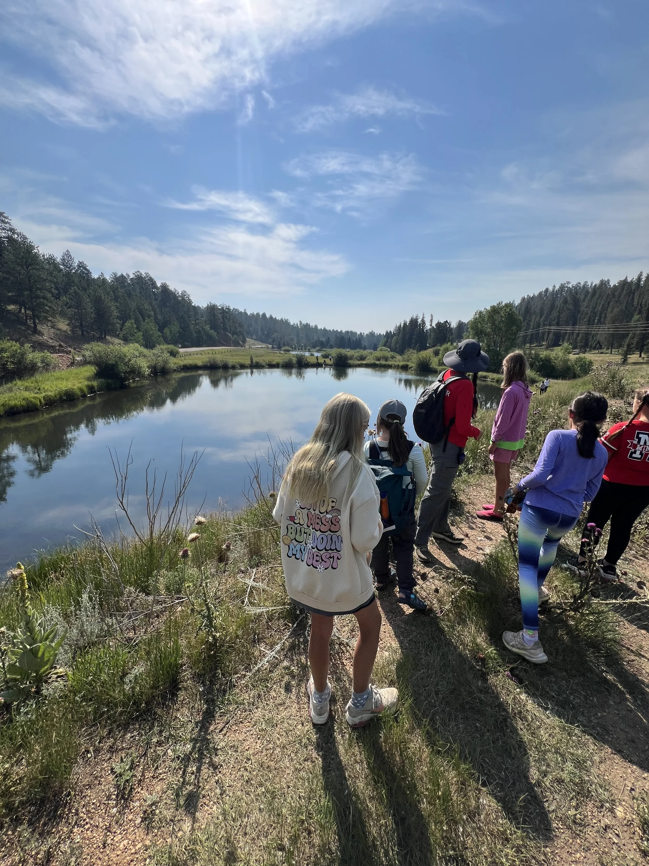 Group of children and adults hiking along a trail by a river in a forested area on a sunny day.
