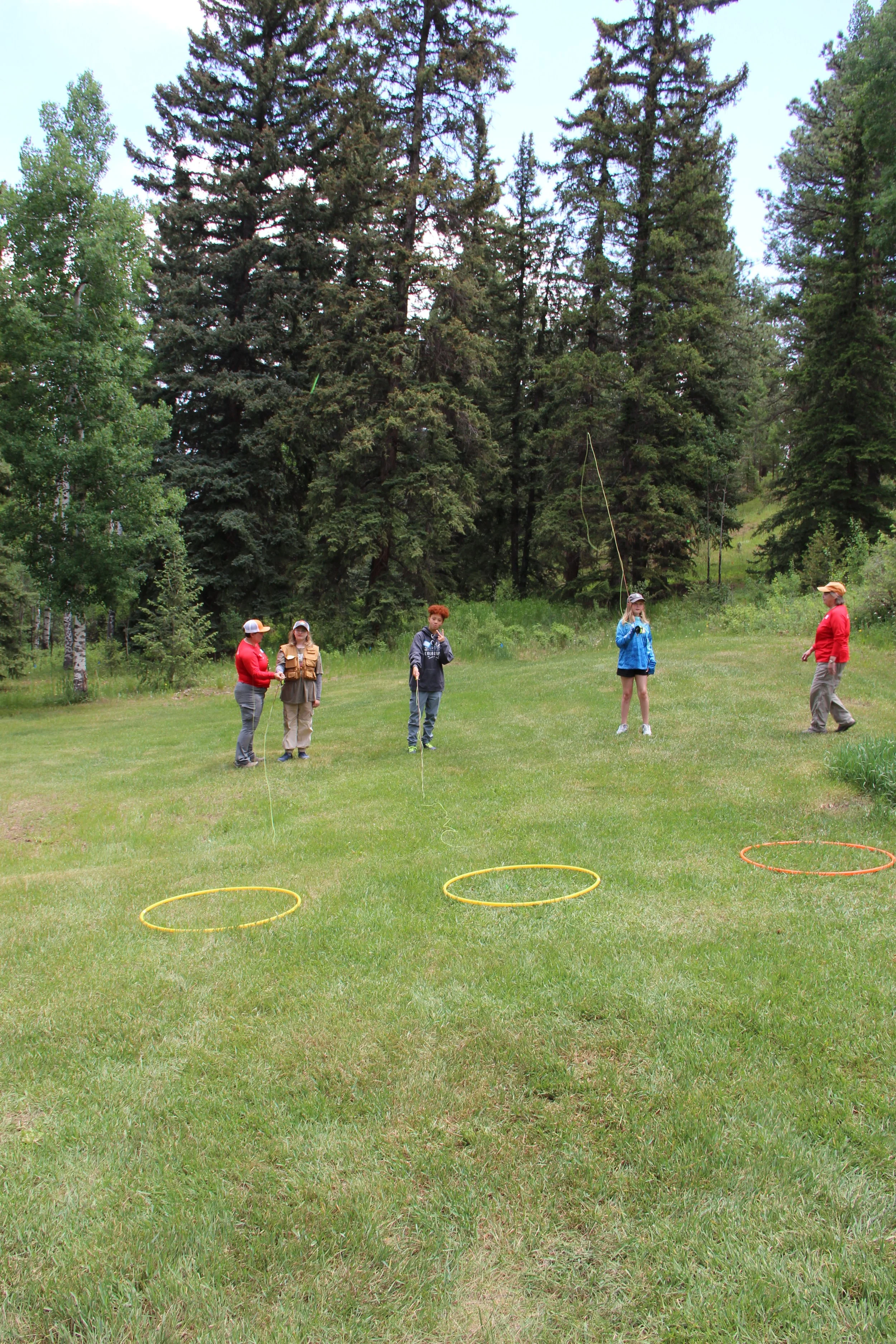 Five people outdoors on a grassy field, preparing for a game or activity with three hula hoops on the ground, surrounded by trees.