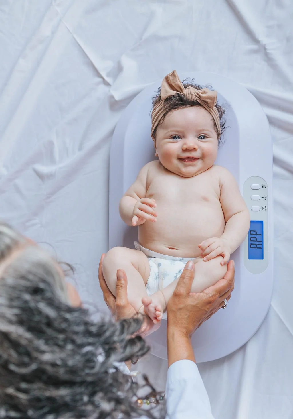 A smiling baby looks at the camera while a Lactation Consultant with brown skin and gray curls gently holds the baby's legs on a scale.