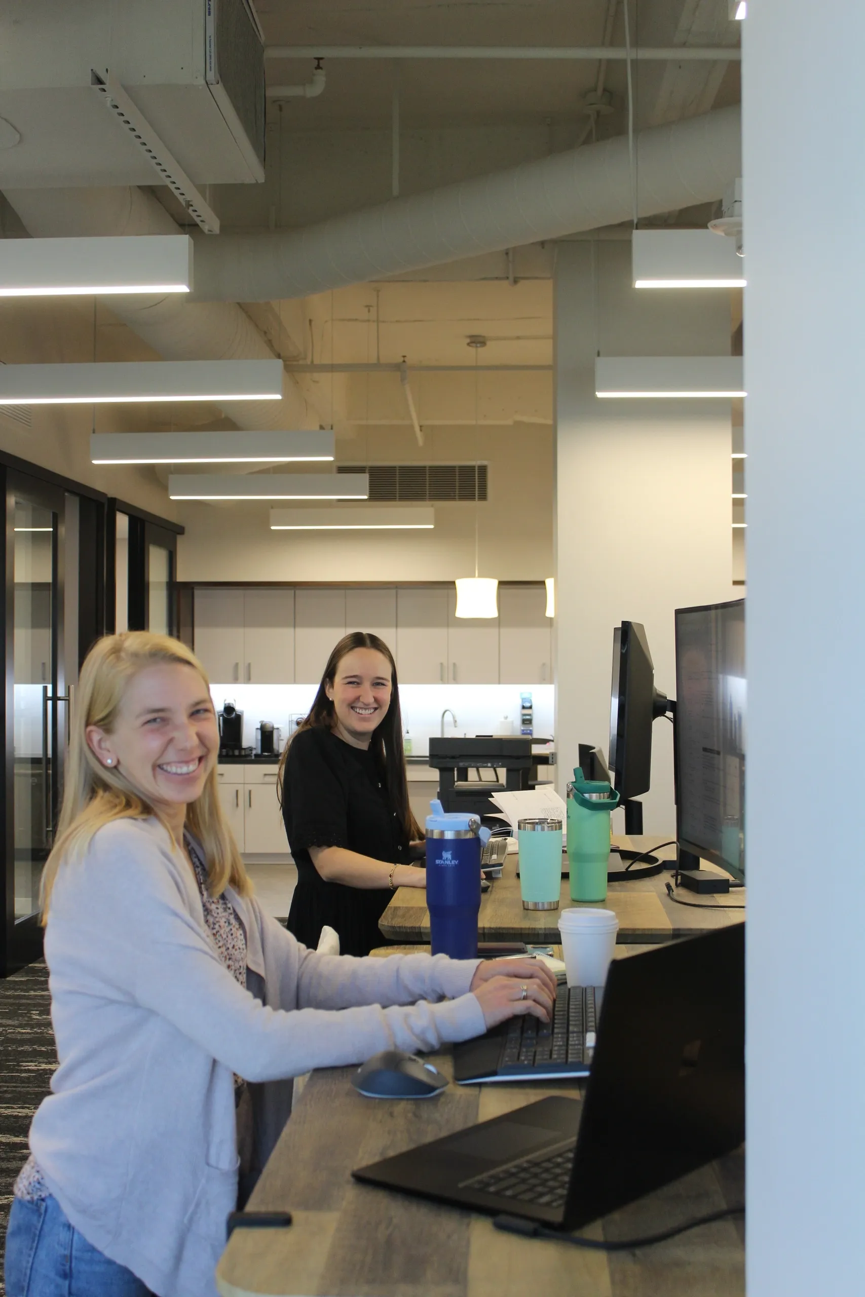 Two women smiling and working at a shared desk with computers in an office setting.