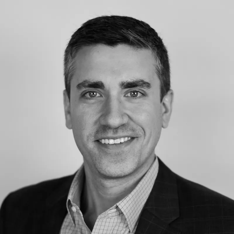 Black and white professional headshot of a man with short dark hair, wearing a checkered shirt and a dark blazer, smiling at the camera.