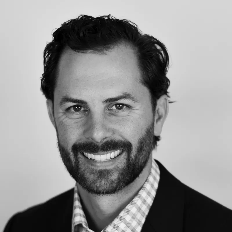 Black and white headshot of a man with dark curly hair, beard, and a checkered shirt, smiling at the camera.