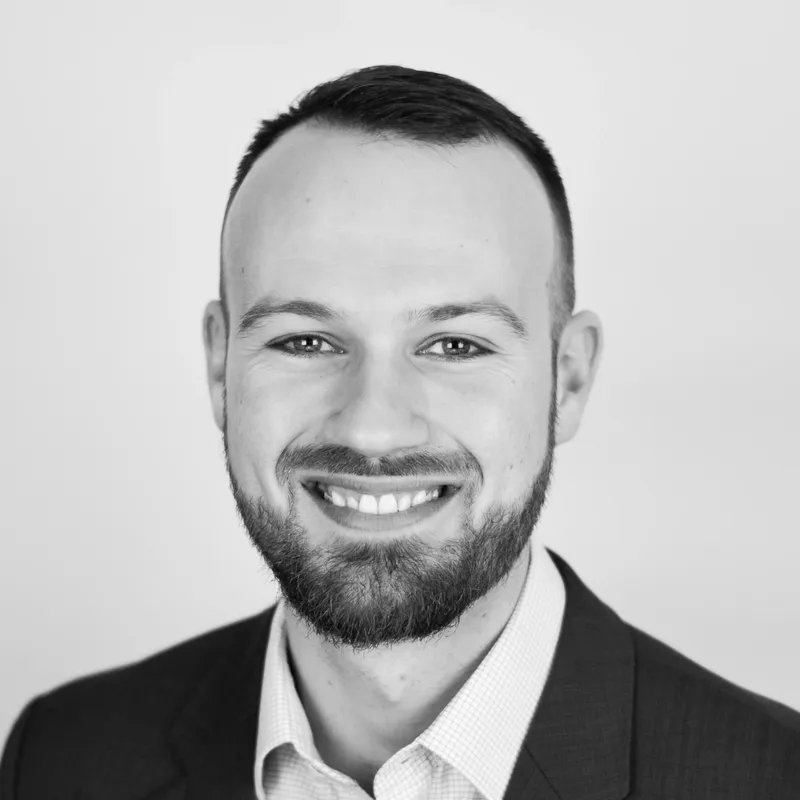 Black and white professional headshot of a smiling man with short hair, a beard, and suit jacket against a plain light background.