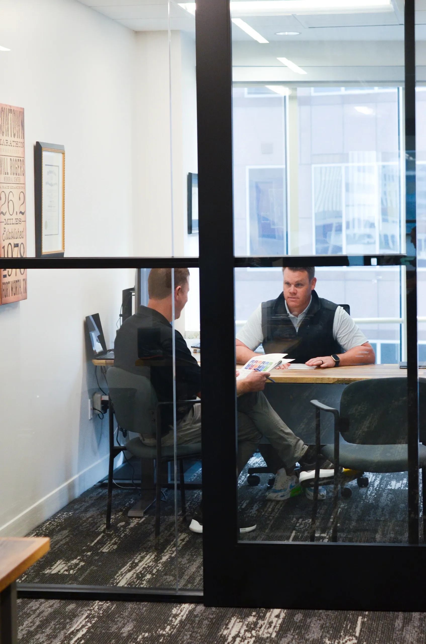 Two men sitting across from each other at a desk in a glass-enclosed office, engaged in a conversation, with one holding a pamphlet or brochure.