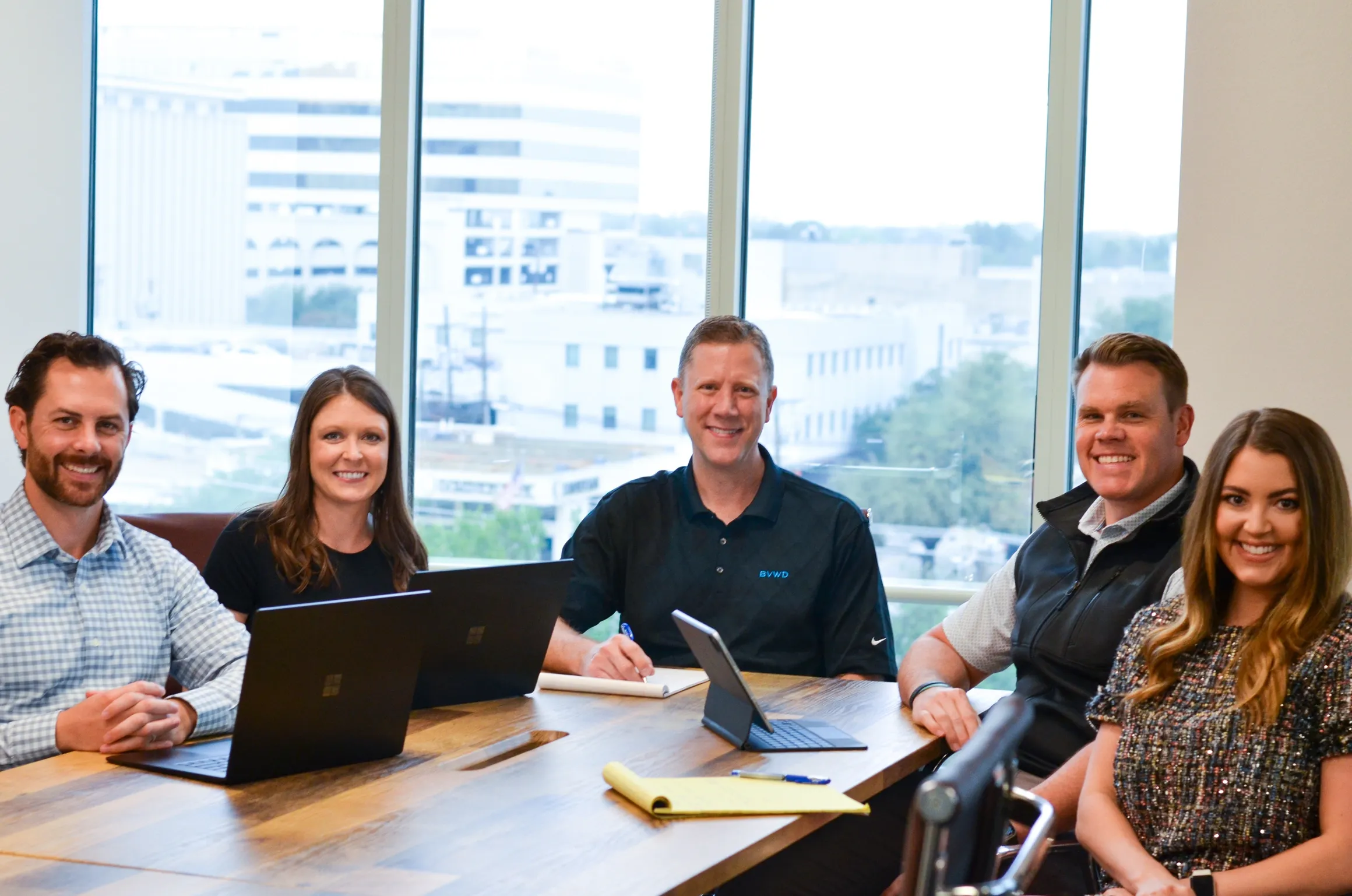 Five people sitting at a conference table in a modern office with large windows, smiling at the camera.