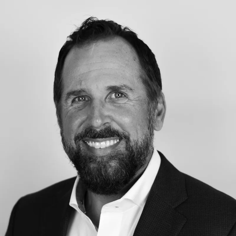 Black and white portrait of a smiling man with a beard, wearing a suit jacket and white shirt, against a plain background.