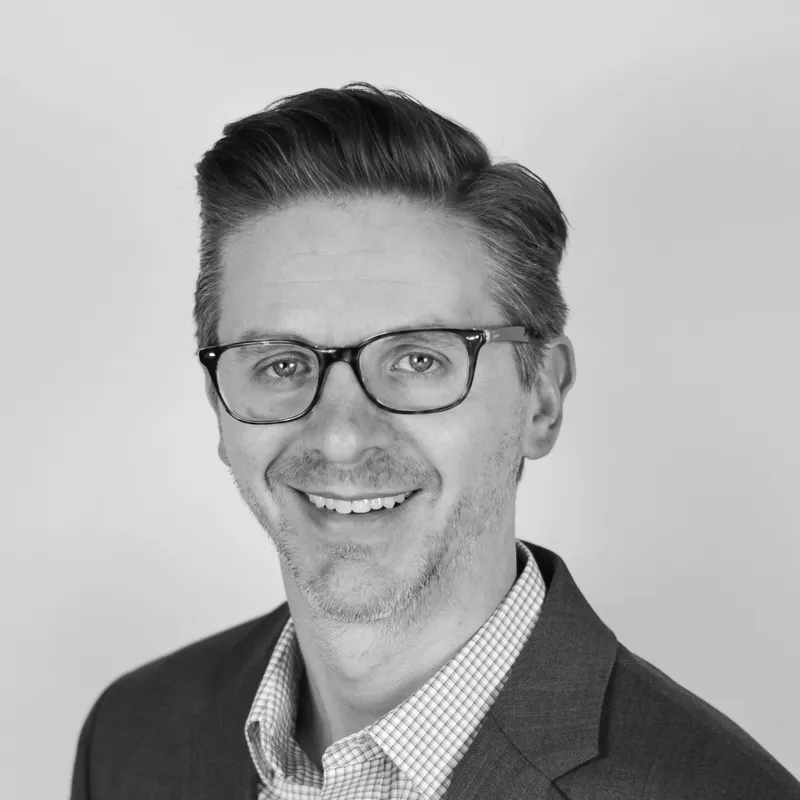 Black and white professional headshot of a man in a suit and glasses, smiling, with styled hair and a background with a gradient from light to darker gray.