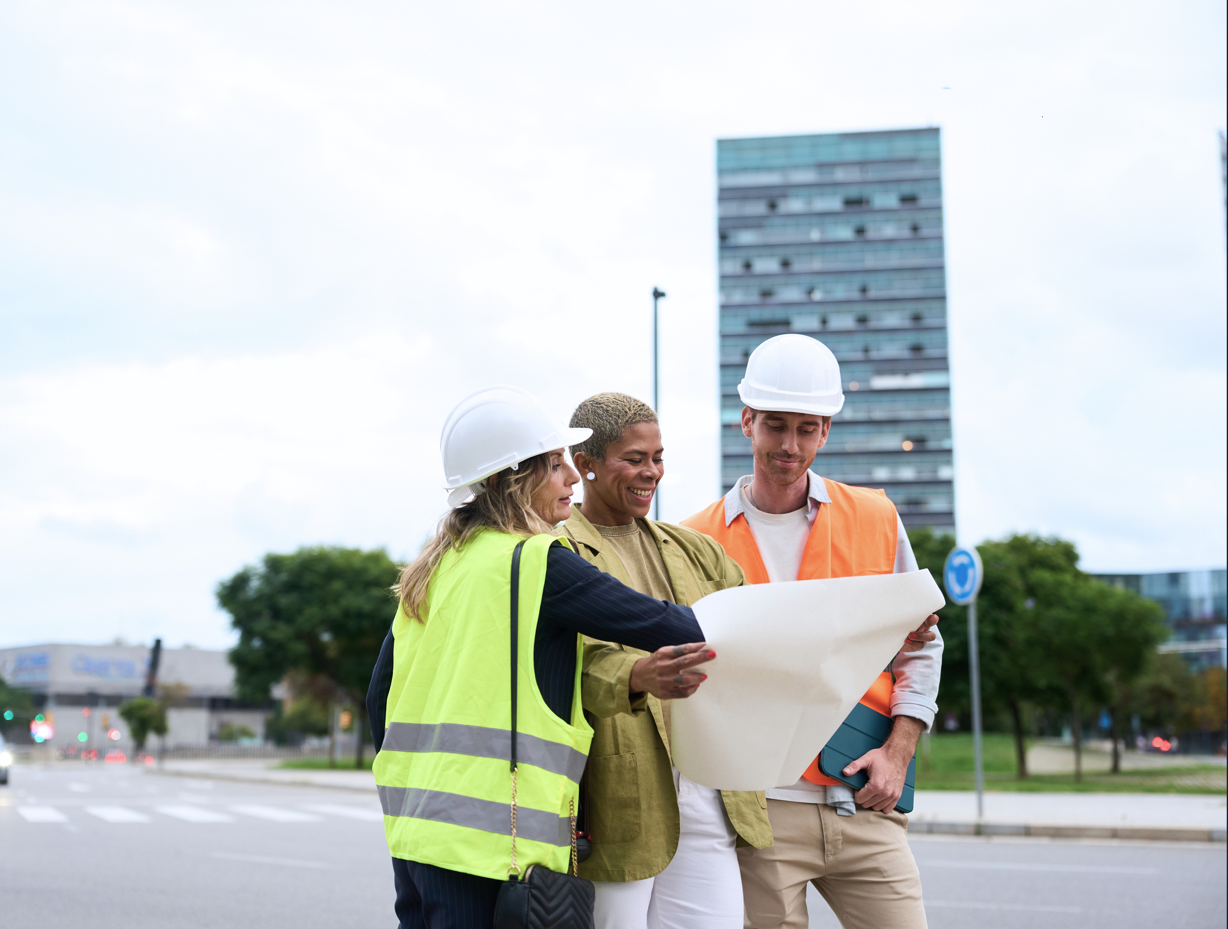 Three construction professionals, two women and one man, stand outdoors wearing safety helmets and vests, reviewing blueprints with a tall building and cityscape in the background.