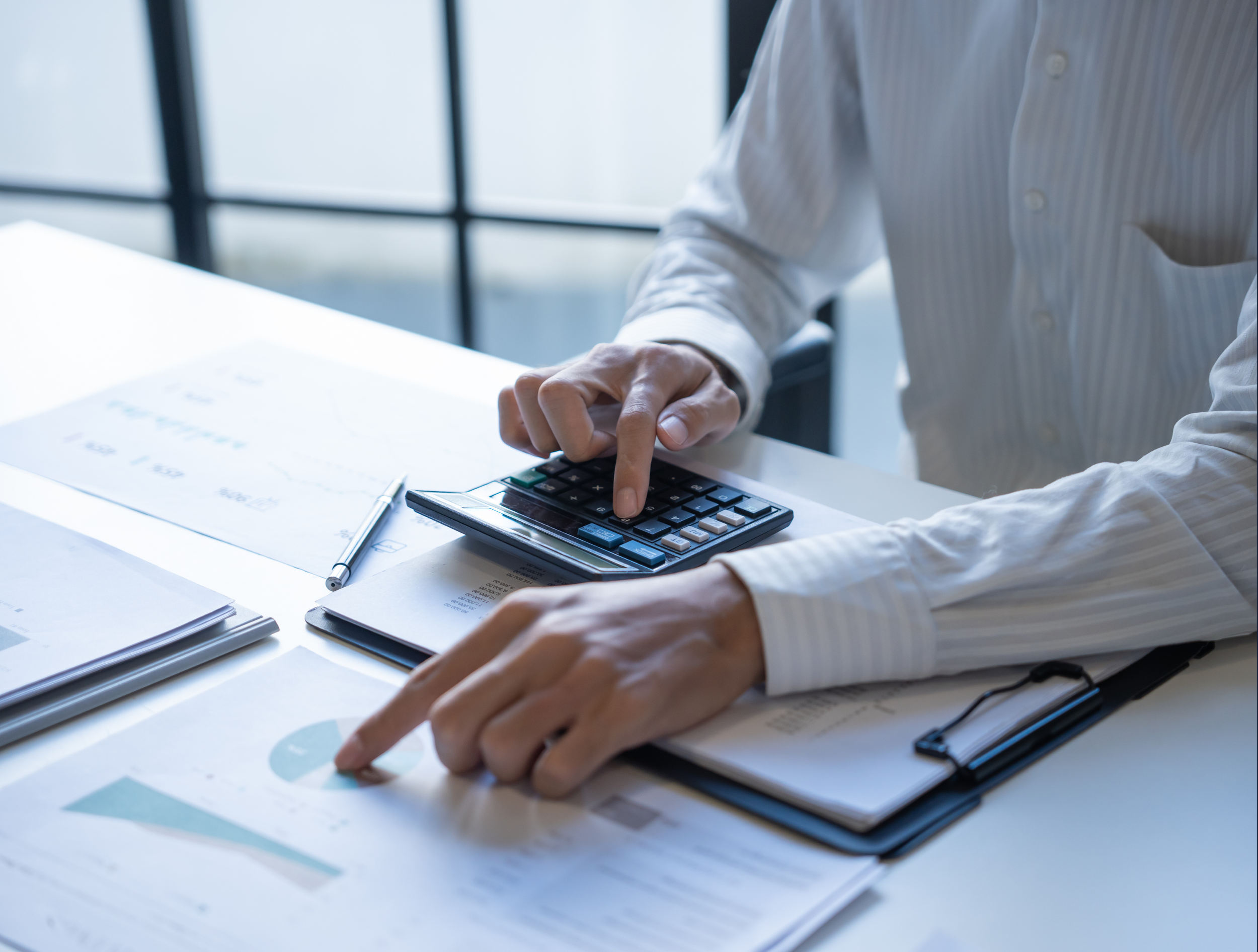 Person using a calculator and pointing to financial documents on a desk with a pen.