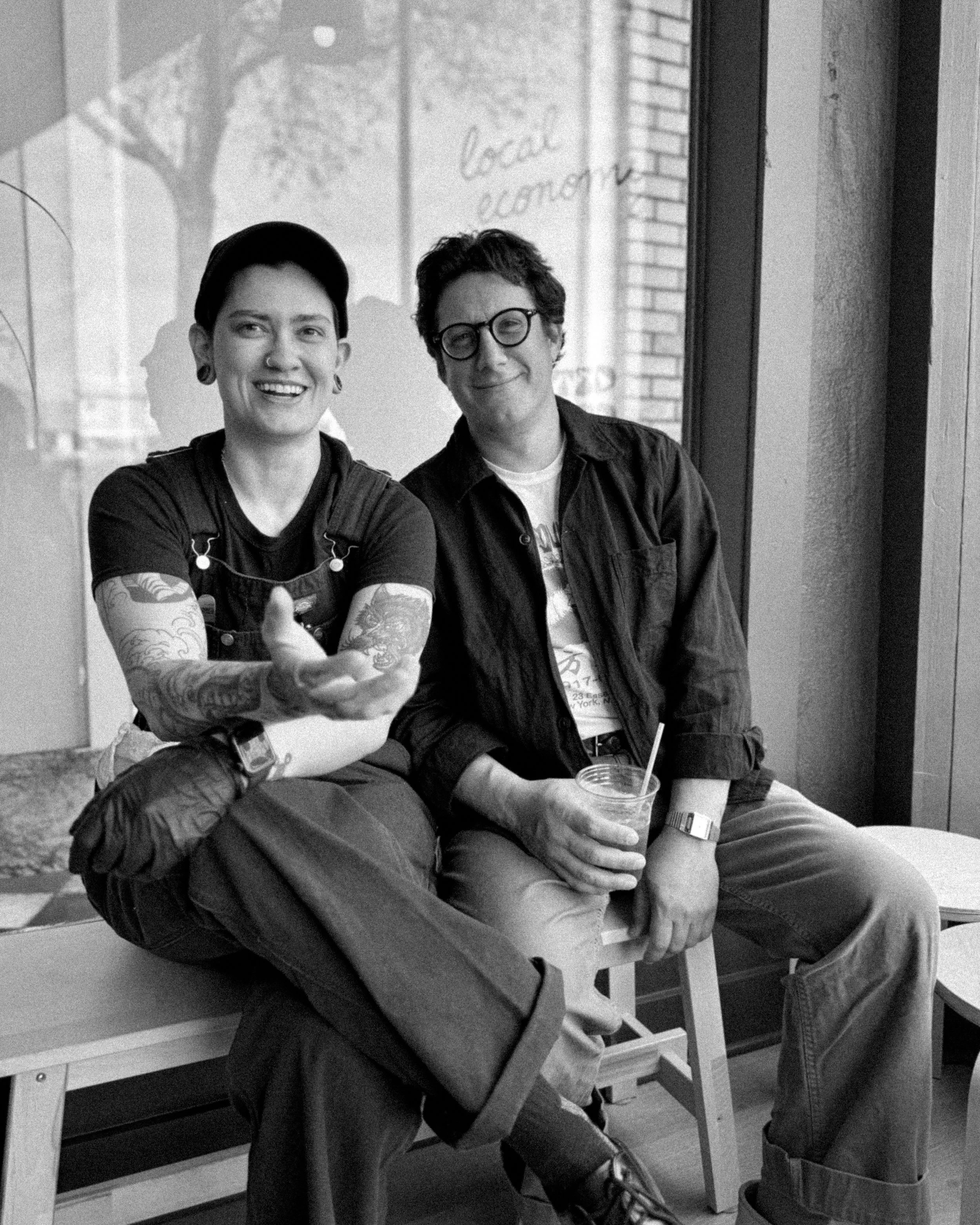 Two people sitting inside a cafe near a large window, smiling and looking at the camera. One person with tattoos and a black cap is gesturing with their hand, while the other person with glasses is holding a drink with a straw.