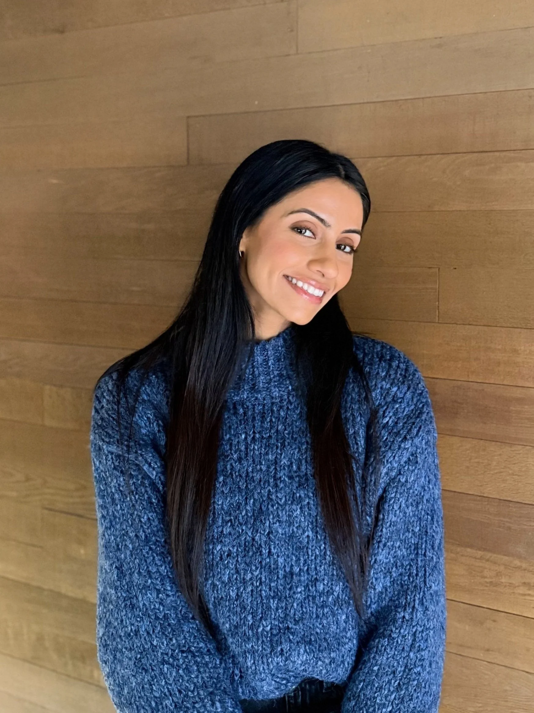 A woman with long dark hair, smiling, wearing a blue knit sweater, standing against a wooden wall.