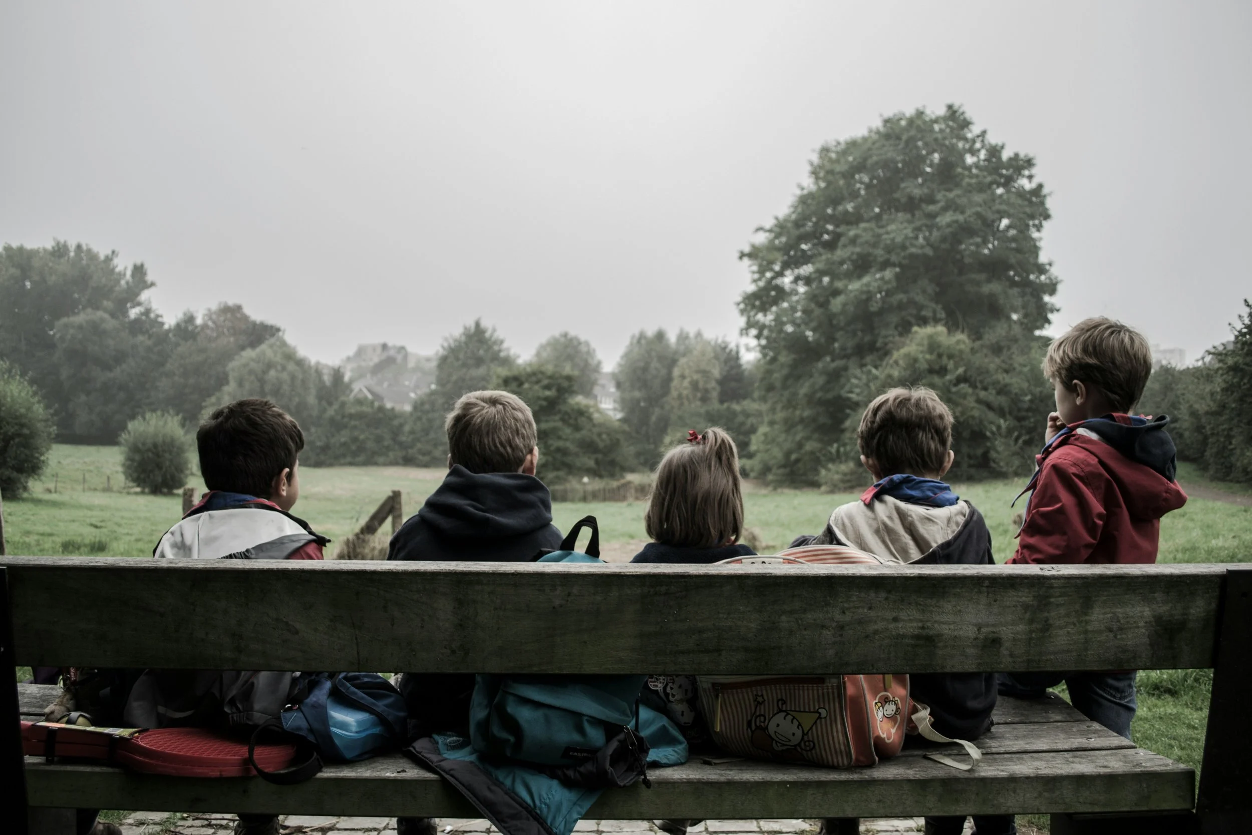 Six children sitting on a wooden park bench, looking at a lush green field with trees, under an overcast sky.