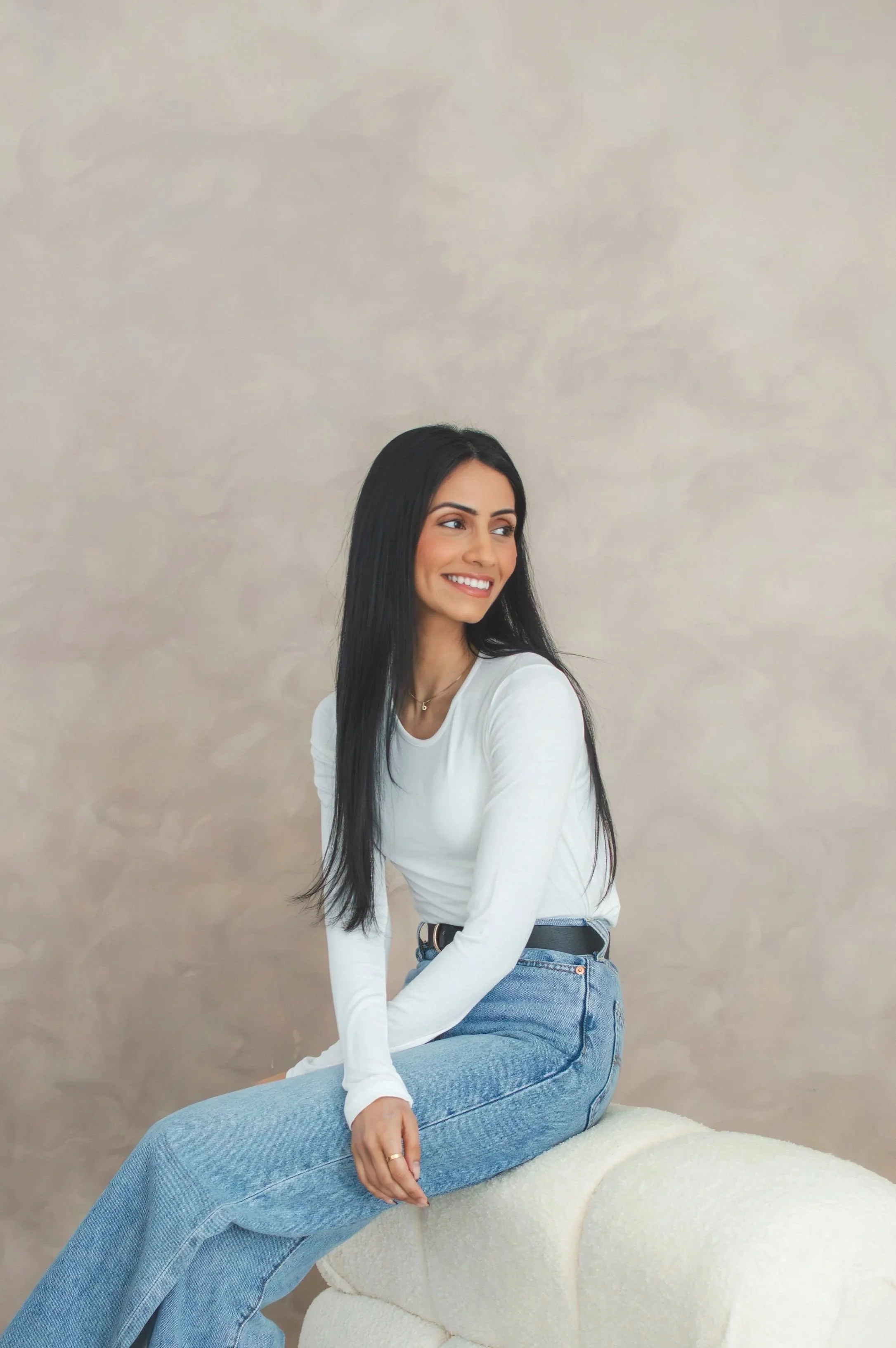 A young woman with long black hair, wearing a white long-sleeve shirt and blue jeans, sitting on a white cushioned stool against a beige background, smiling and looking to her left.
