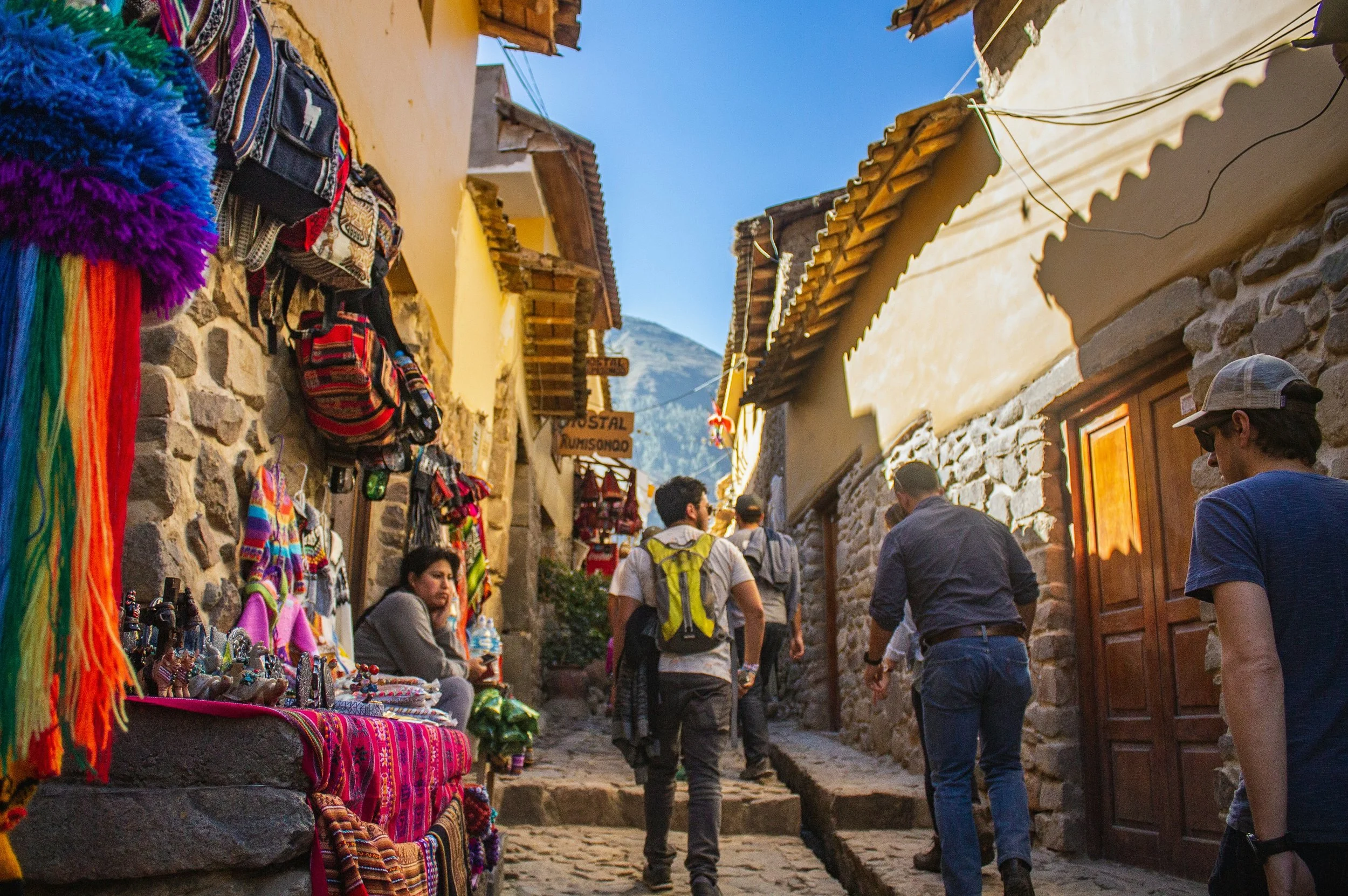 A narrow cobblestone street in a tourist town with shops selling colorful textiles and souvenirs, with several visitors walking and browsing.