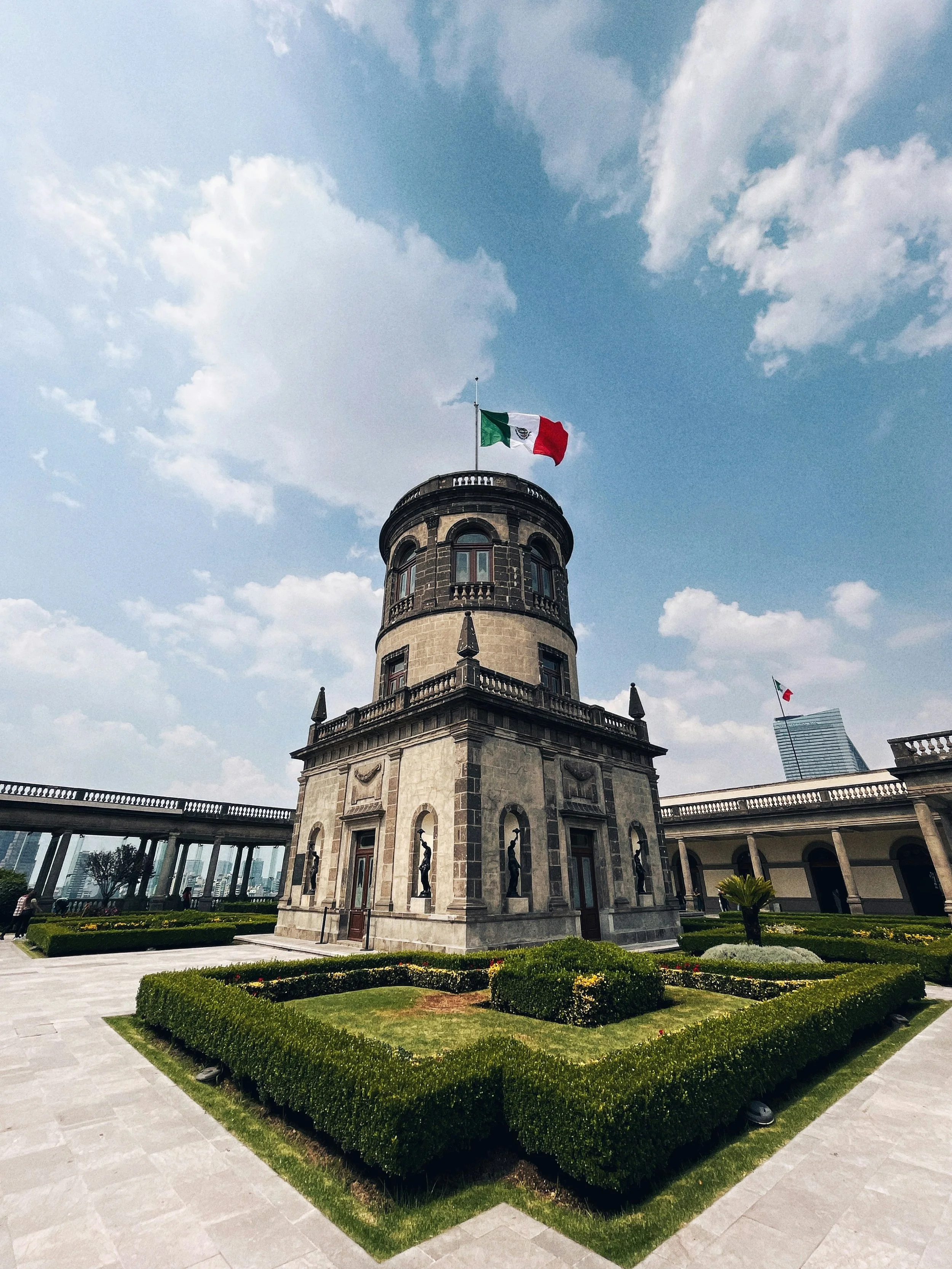 Historical tower with Mexican flags on top, surrounded by manicured gardens and a clear blue sky with clouds.