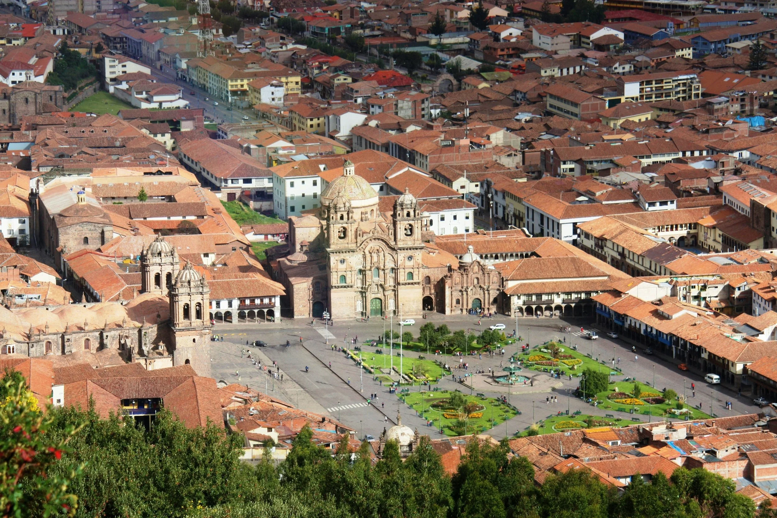 Aerial view of a historic city square with a large cathedral in the center, surrounded by red-tiled roof buildings, cobblestone streets, and well-maintained green gardens with flower beds and fountains.