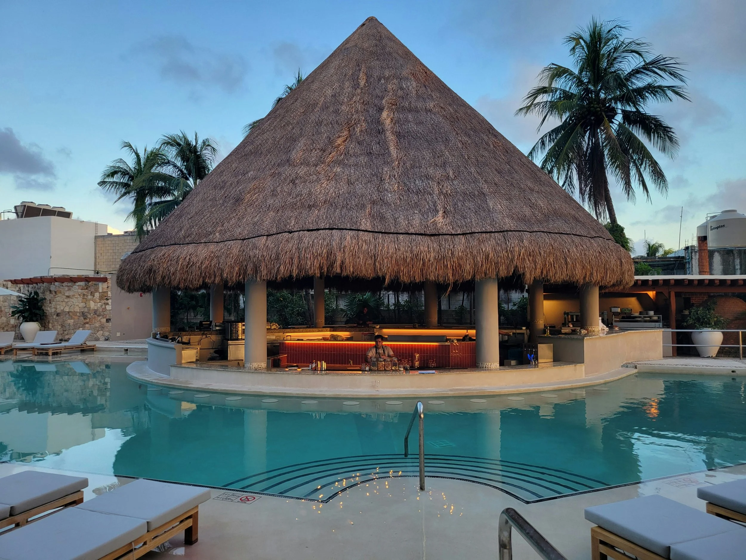 Elegant swimming pool area with a thatched-roof poolside bar, surrounded by palm trees and lounge chairs at sunset.