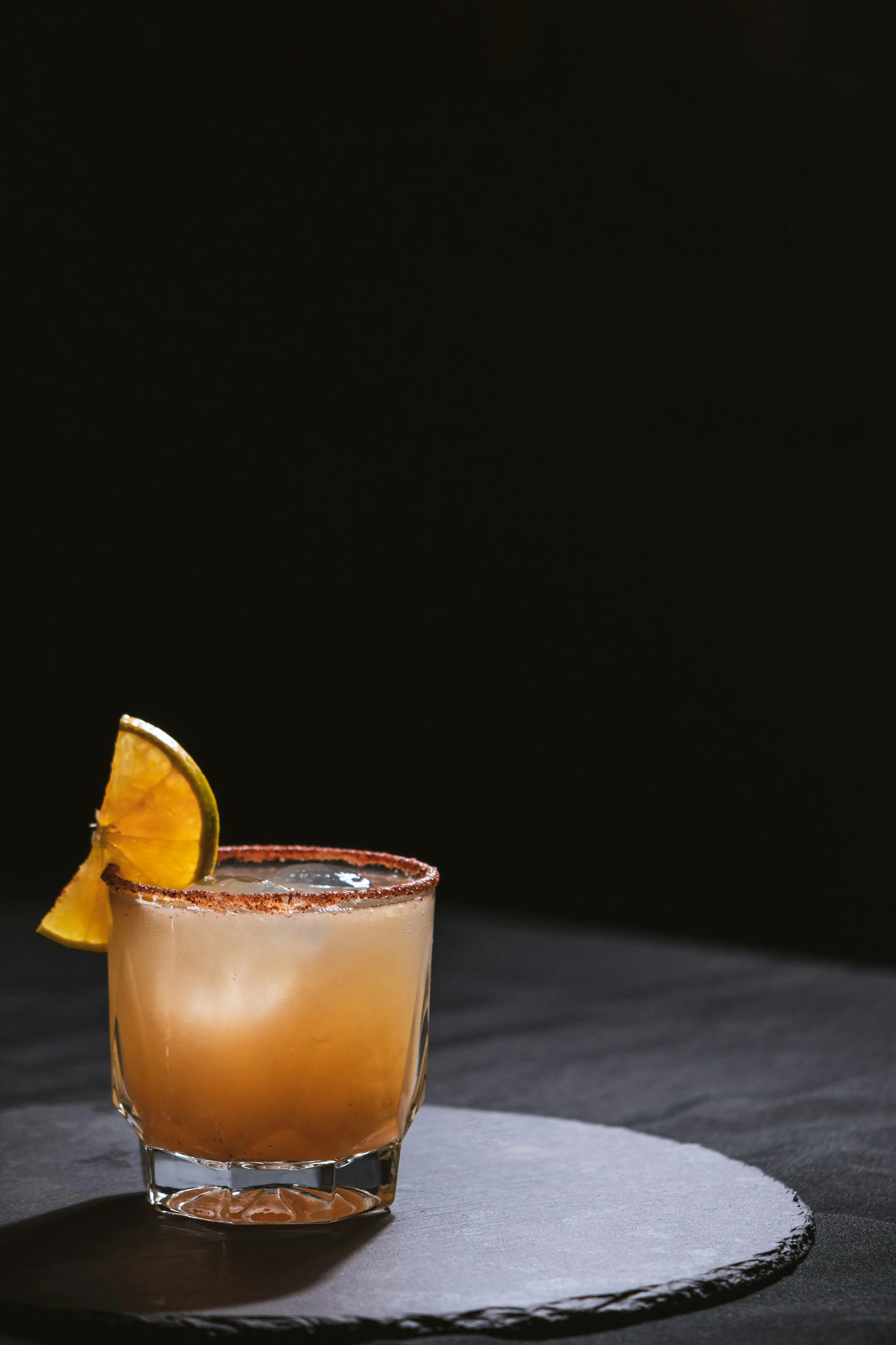 A cocktail in a short glass with a salted rim, garnished with a lemon wedge, placed on a dark surface with a black background.
