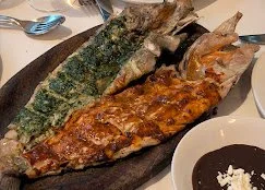 Two whole grilled fish served on a black plate with sauce and side dishes, with utensils nearby.