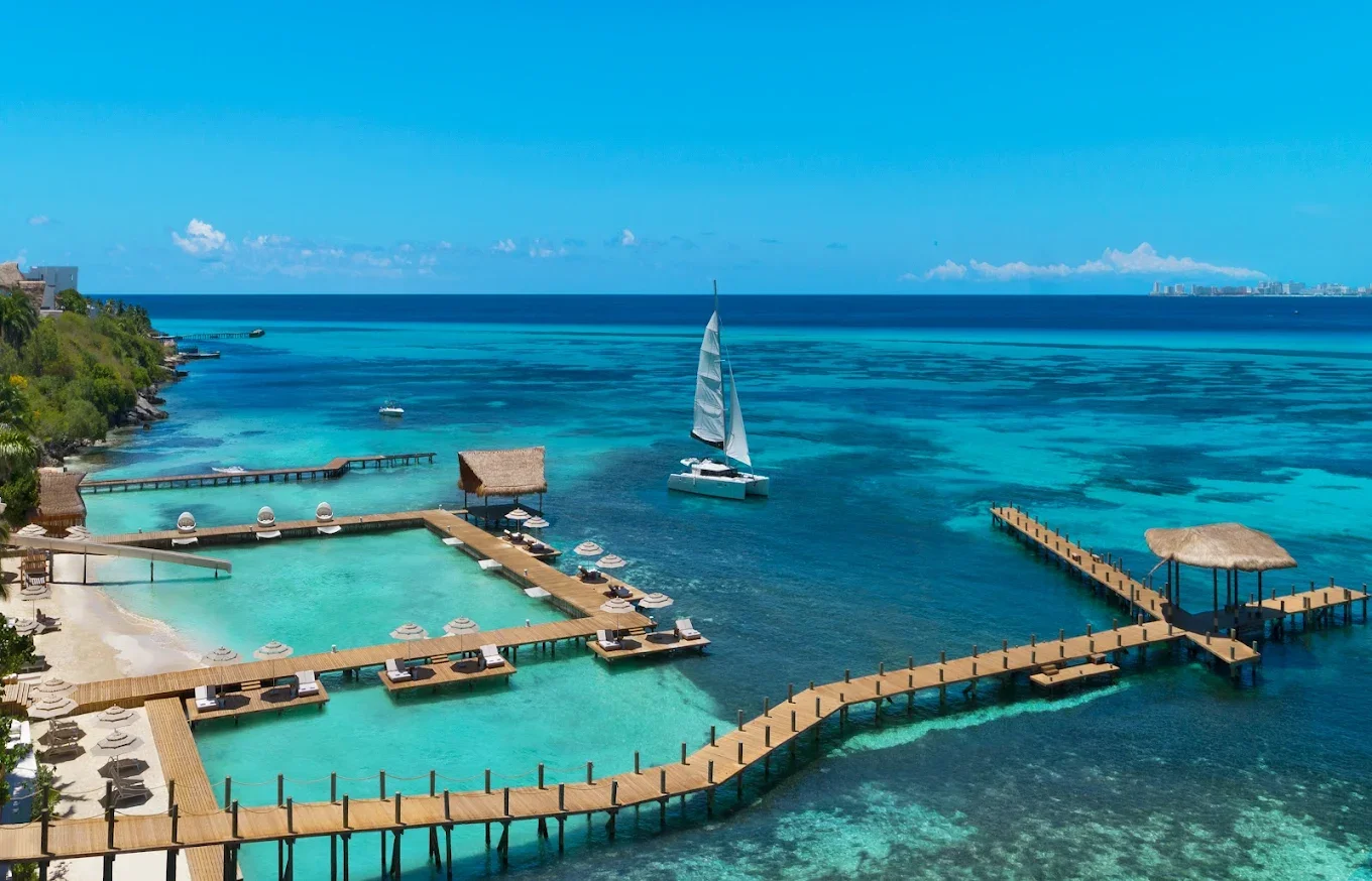 Overwater resort with wooden decks, swimming pools, thatched-roof pavilions, and a sailboat on turquoise ocean waters under a clear blue sky.