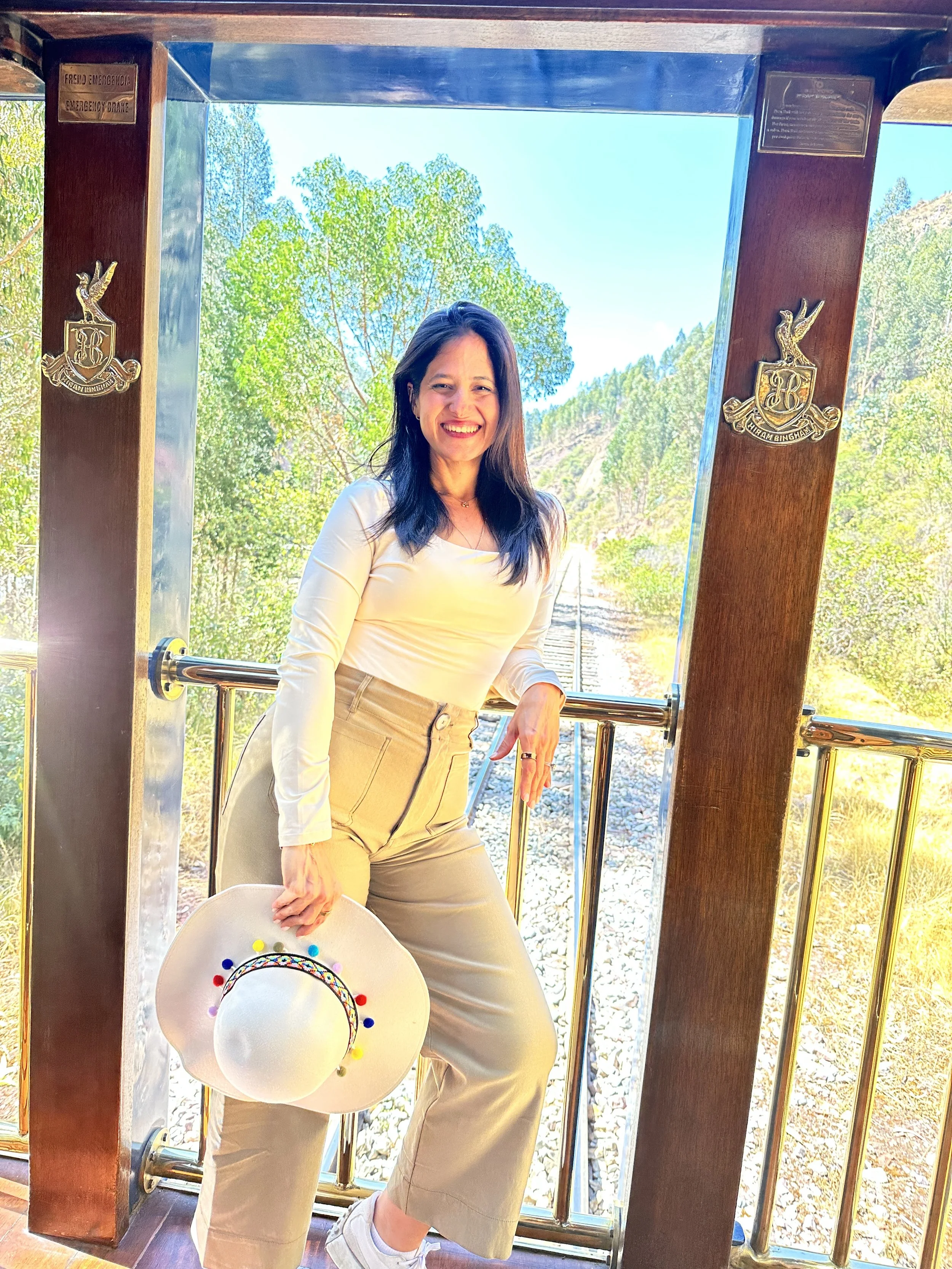 Woman smiling and holding a colorful hat, standing on a train station platform with greenery and train tracks in the background.